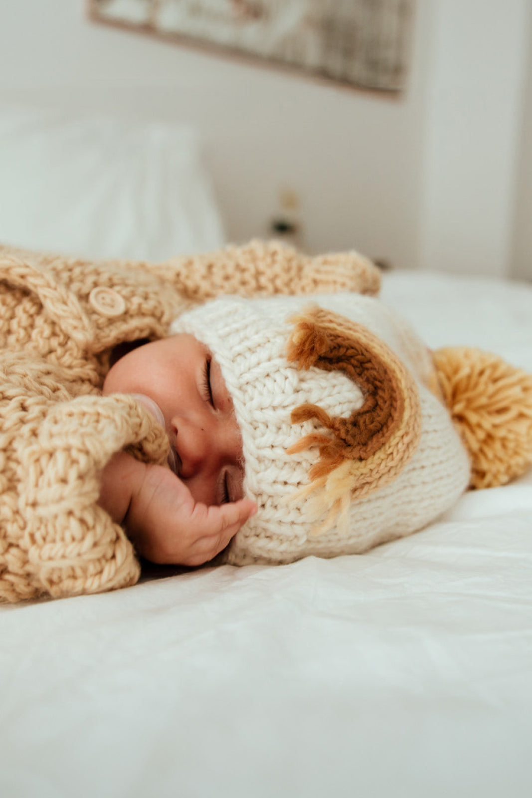 Baby sleeping peacefully on a bed, wearing a cozy knit sweater and a cute hat with a pom-pom.