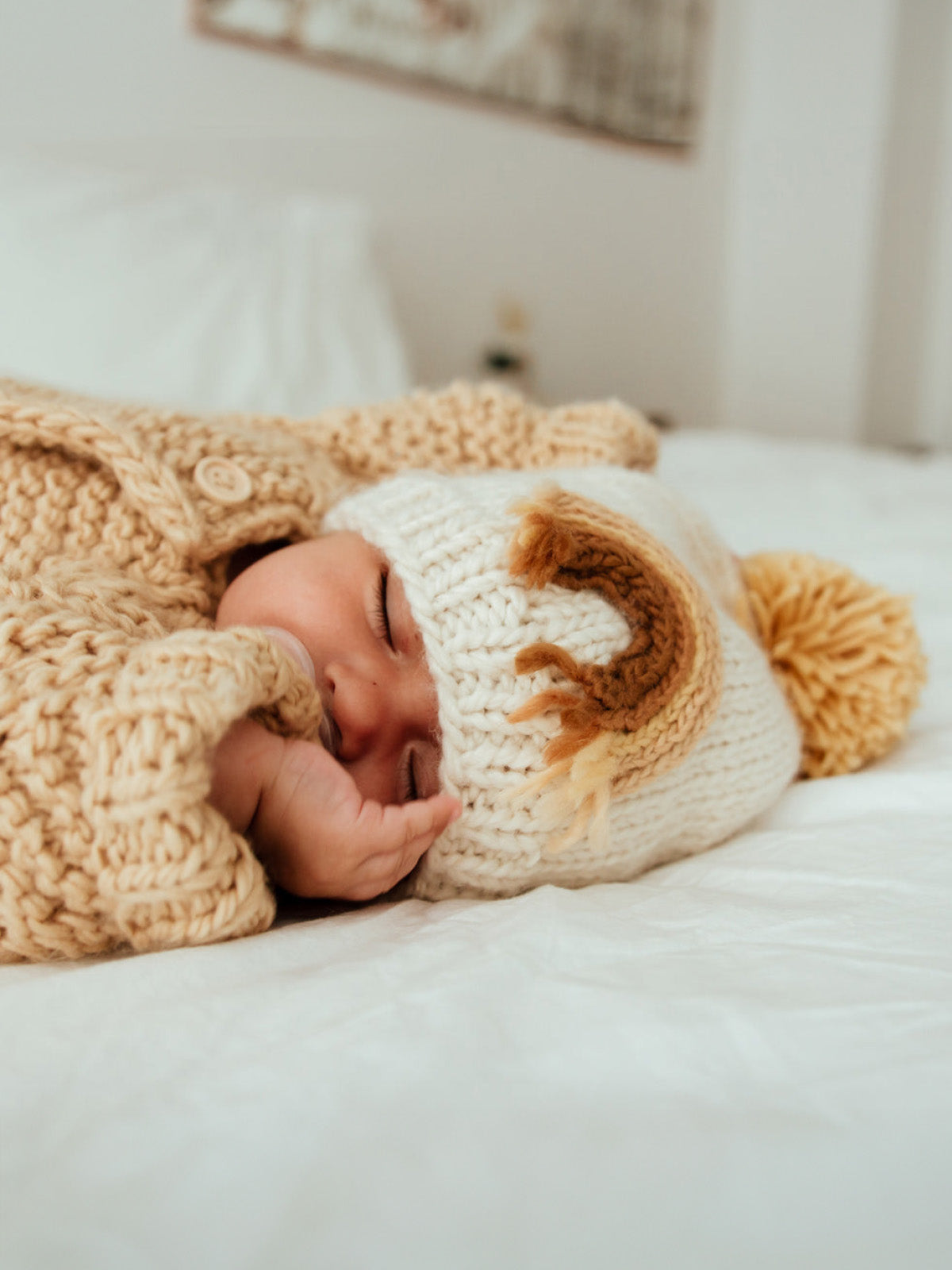 Baby sleeping peacefully on a bed, wearing a cozy knit sweater and a cute hat with a pom-pom.