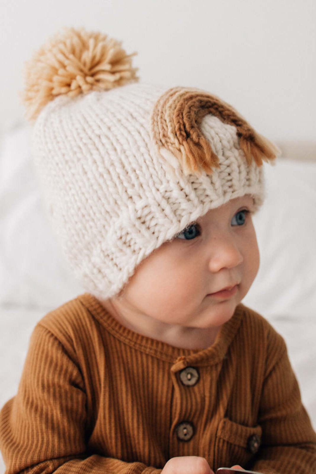 Baby wearing a knotted cream beanie with a pom-pom, sitting on a bed in natural light.