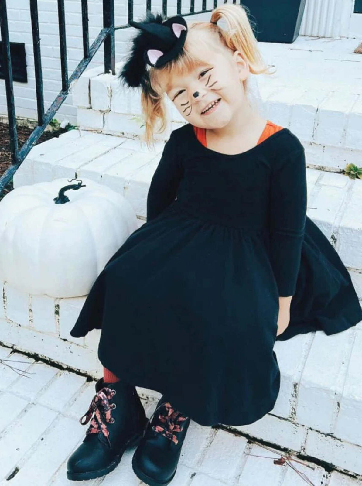 Smiling child in a black cat costume sitting by a white pumpkin on a porch.