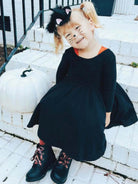 Smiling child in a black cat costume sitting by a white pumpkin on a porch.
