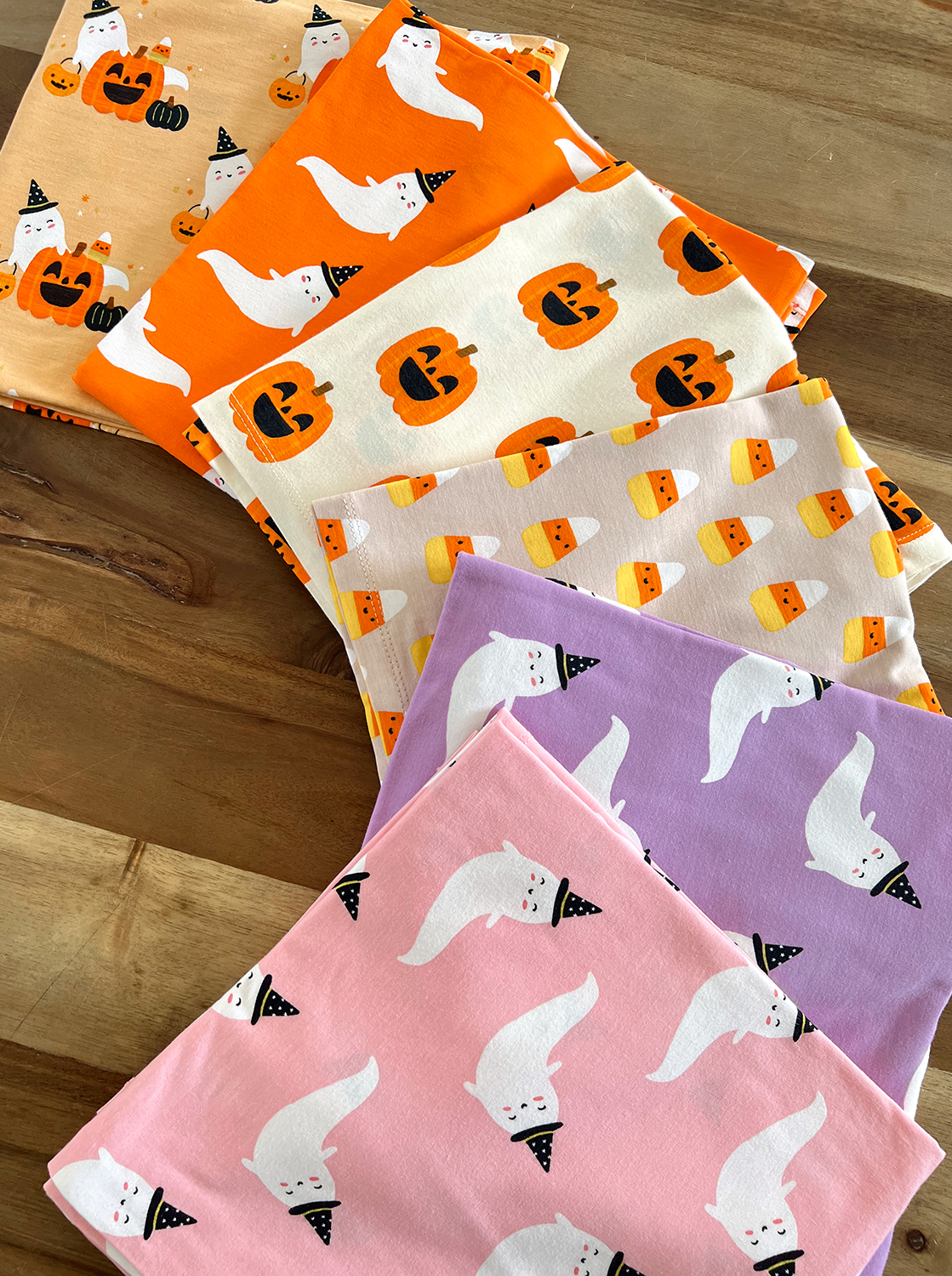 Colorful Halloween-themed fabrics featuring ghosts, pumpkins, and candy corn stacked on a wooden surface.