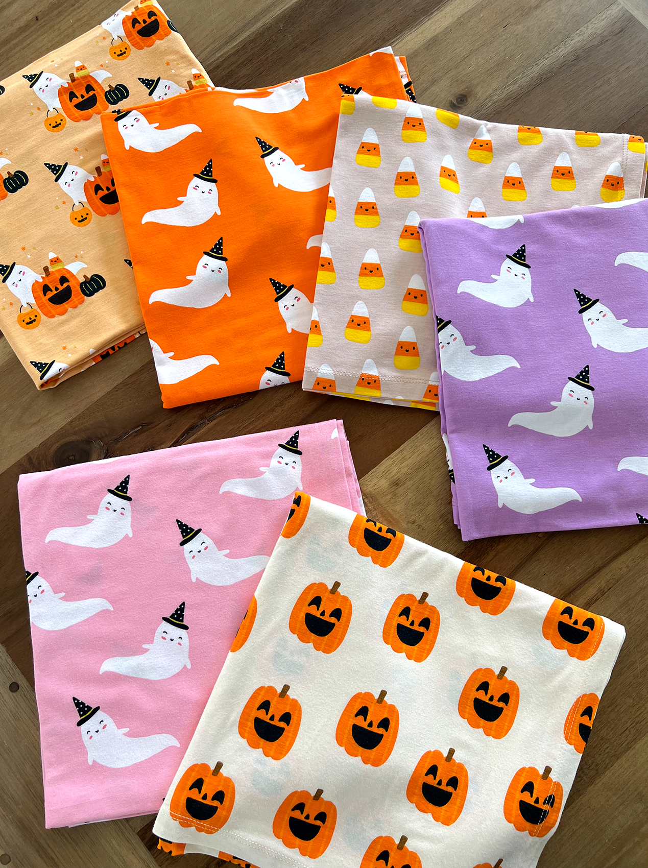 Colorful Halloween-themed fabrics featuring ghosts, pumpkins, and candy corn arranged on a wooden surface.