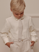 Boy in a cream shirt and beige pants, looking down, with hands in pockets against a neutral background.