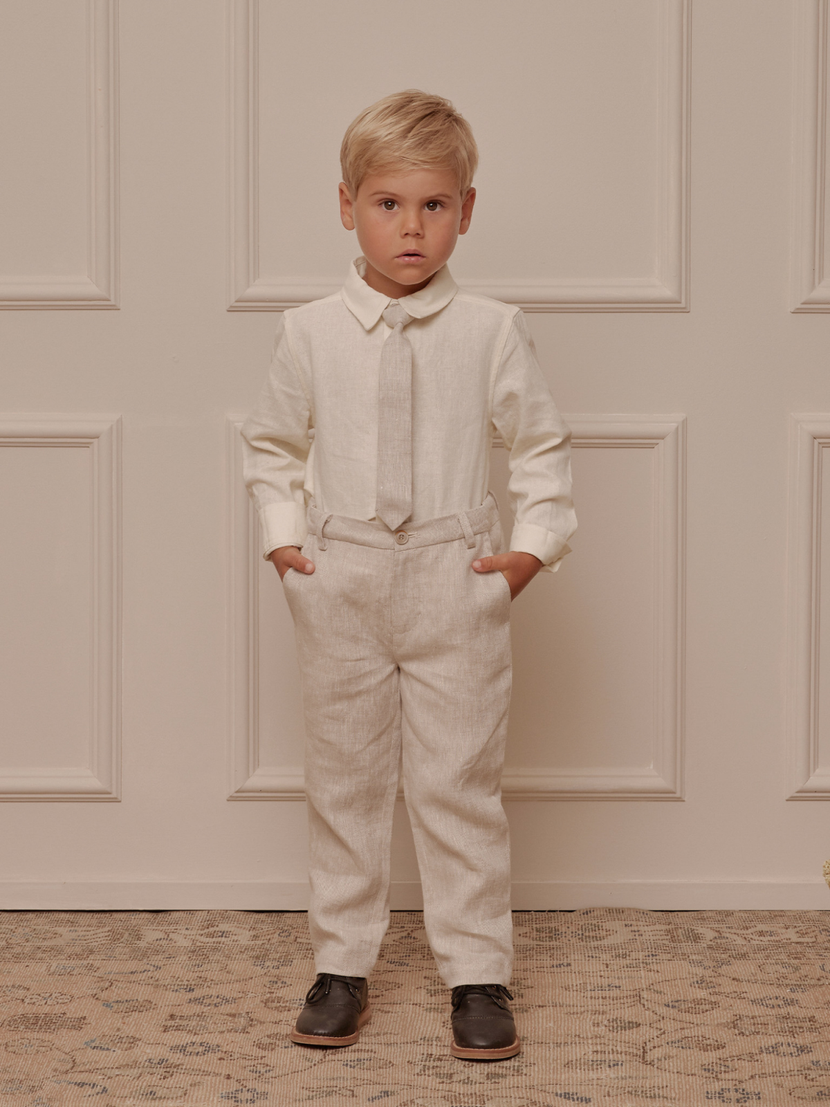 Young boy in light outfit with a tie, standing confidently in front of a textured backdrop.
