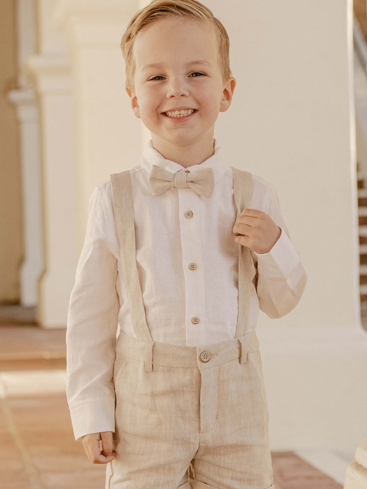 Smiling boy in a beige bow tie and suspenders, dressed in a light shirt and shorts, standing indoors.