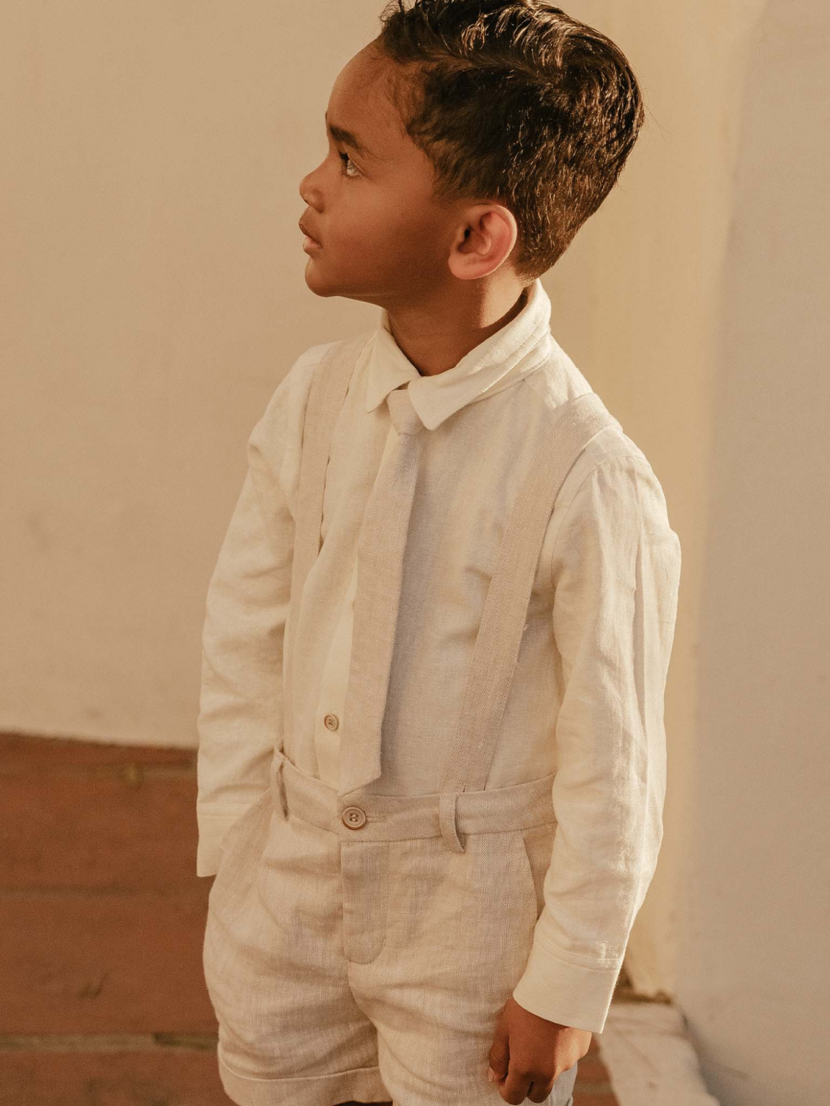 Young boy in a light linen suit and tie, standing against a soft beige background.