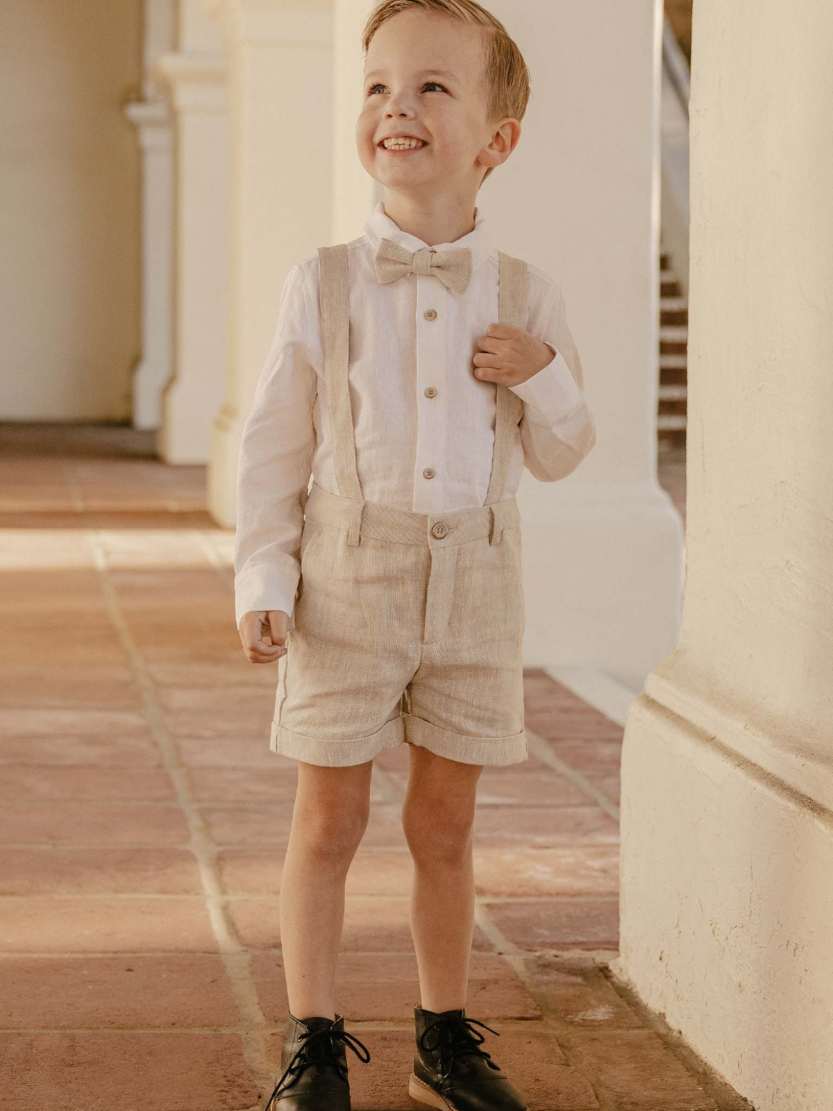 Smiling boy in a stylish outfit with bowtie and suspenders, standing on a tiled floor in a bright setting.