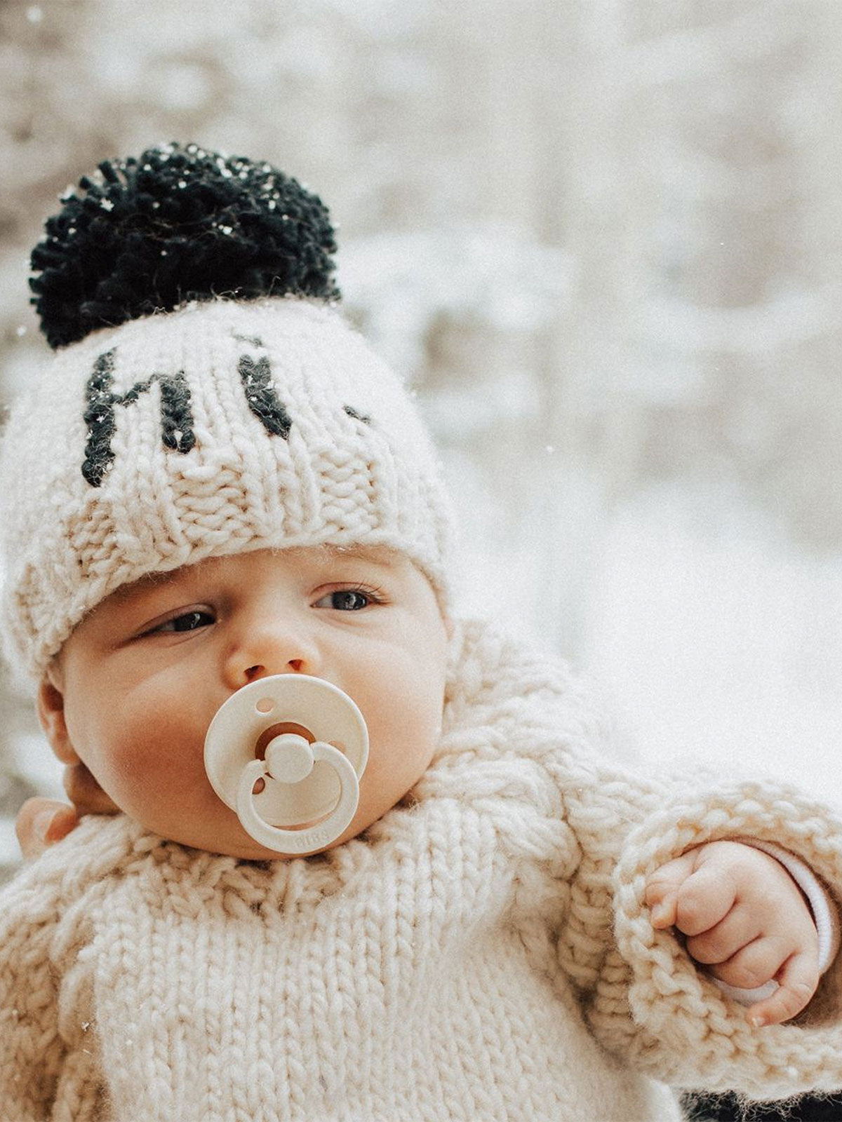 Baby wearing a knitted hat with a pom-pom, holding a pacifier, in a snowy outdoor setting.