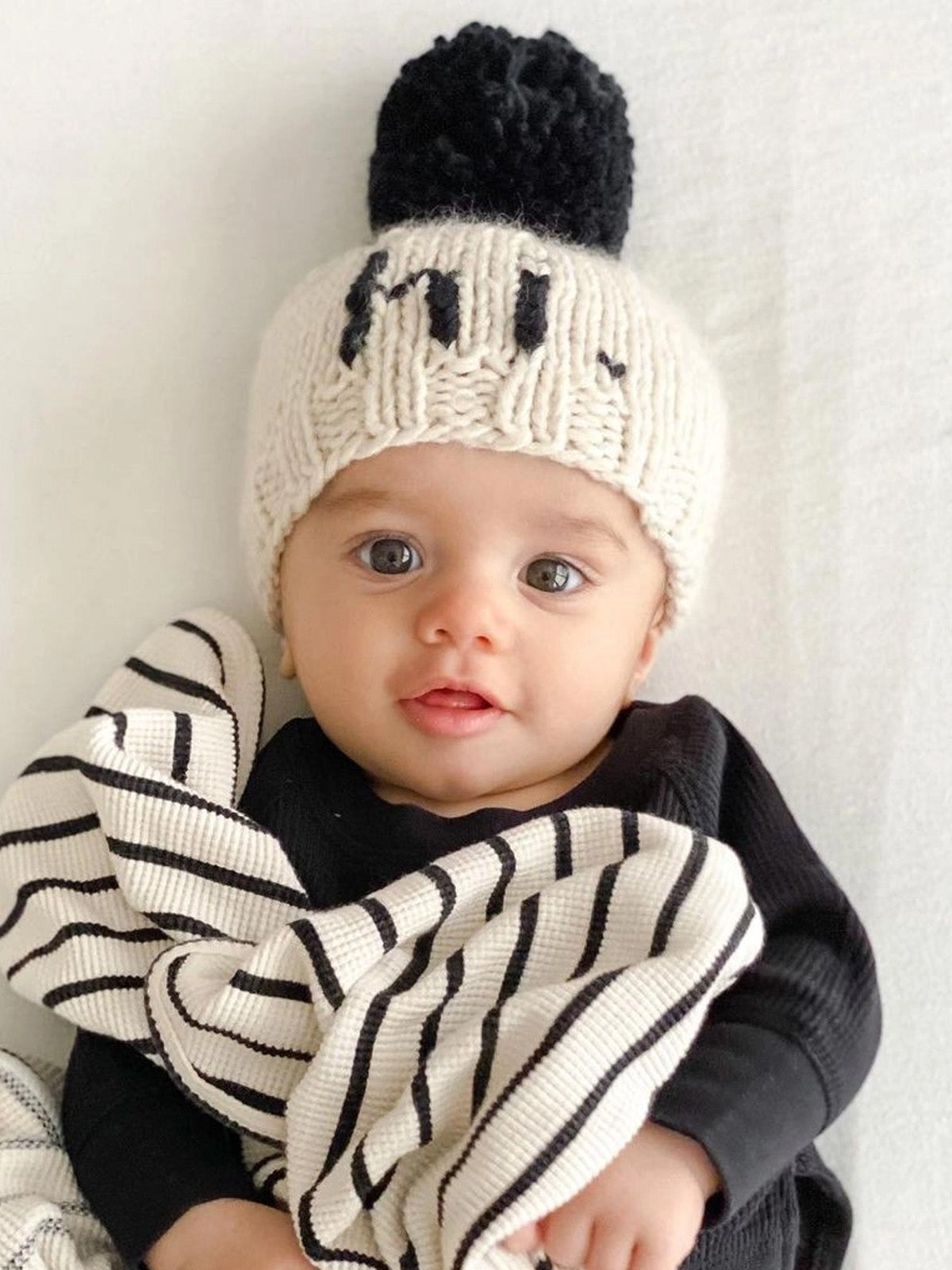 Baby wearing a knitted hat with a black pom-pom, smiling and draped in a striped blanket.