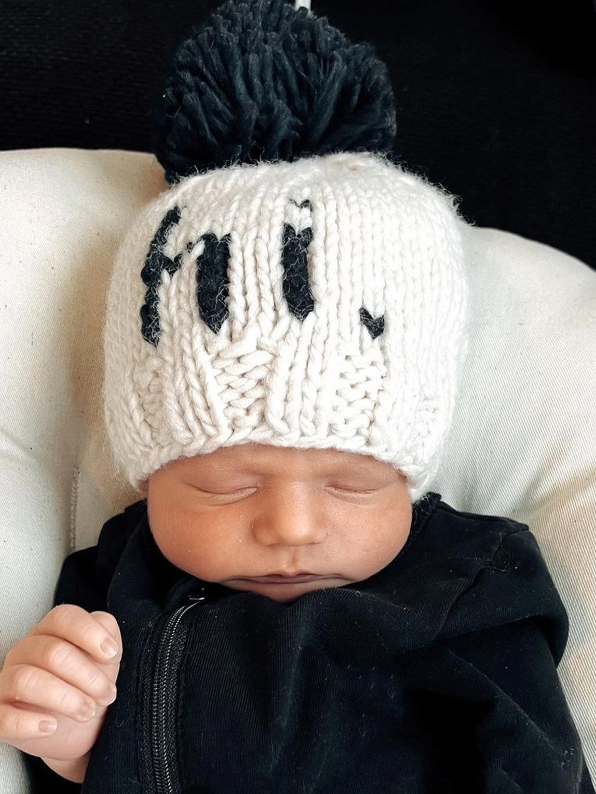 Baby sleeping in a cozy black outfit, wearing a white knitted hat with a black pom-pom and the letters "hi."
