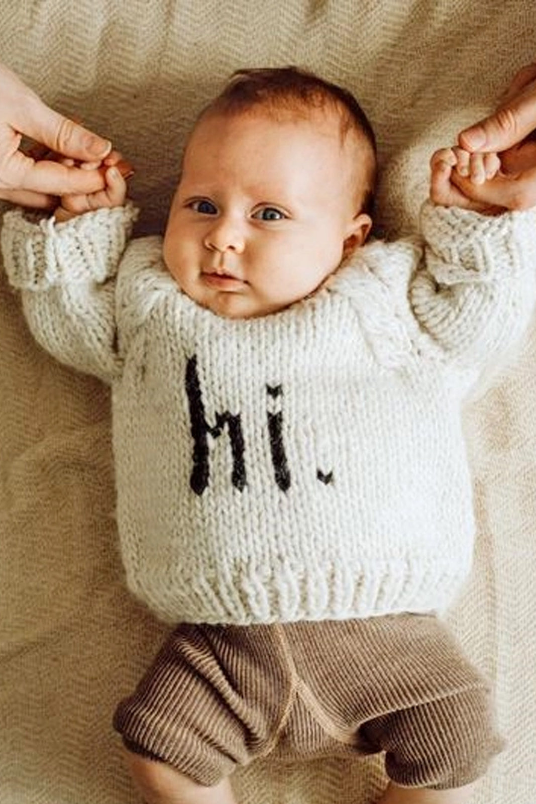 Baby in a cozy knit sweater with "hi" printed, being held by two hands, on a soft textured blanket.
