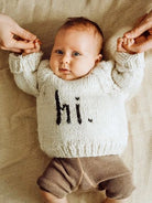 Baby in a cozy knit sweater with "hi" printed, being held by two hands, on a soft textured blanket.