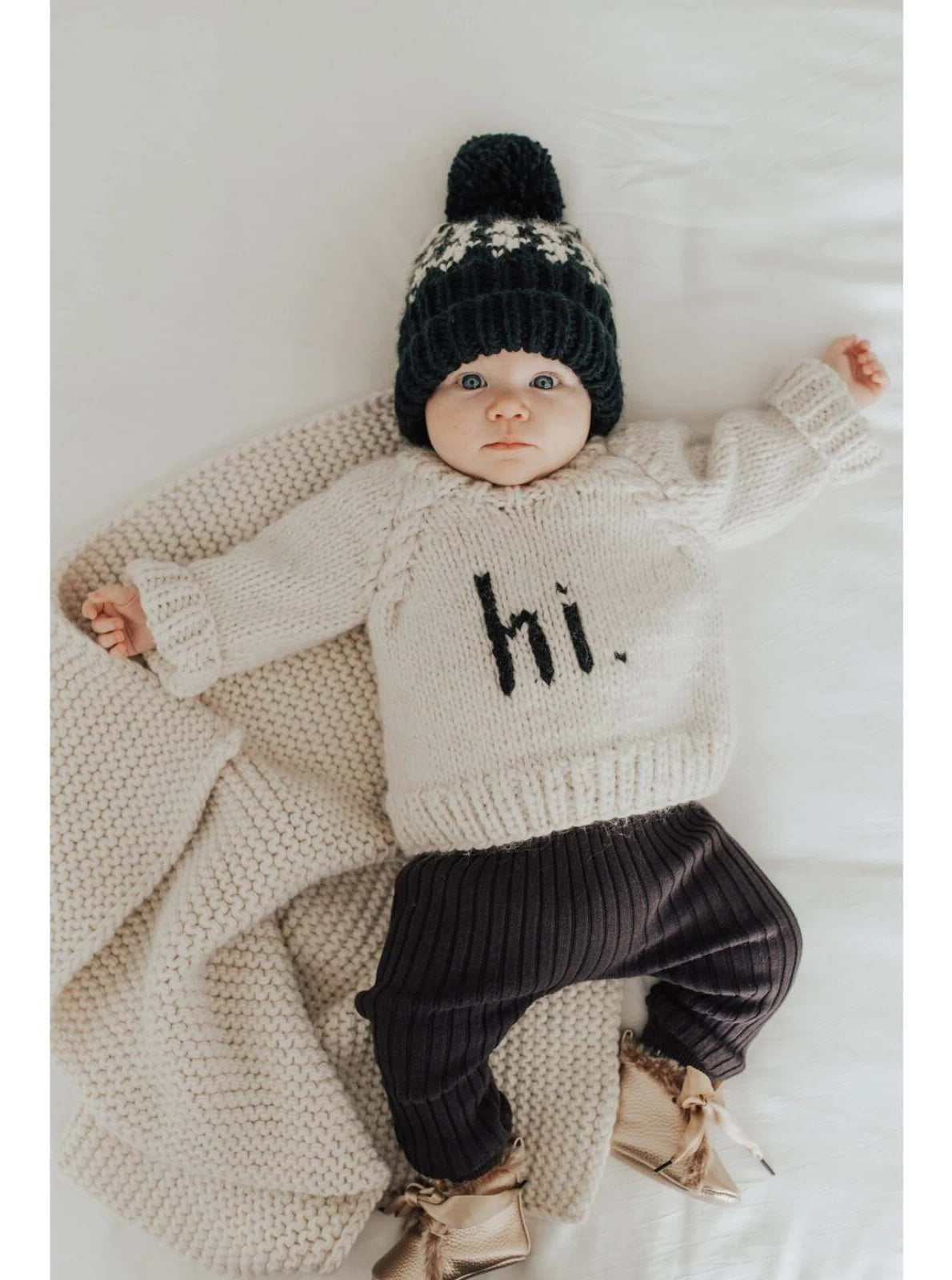 Baby in a knitted sweater with "hi." text, wearing a beanie, lying on a soft blanket.