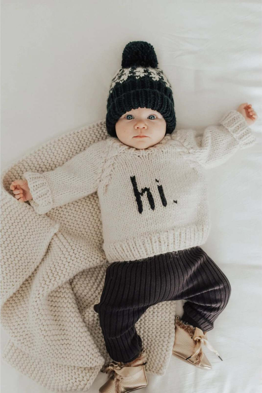 Baby in a knitted sweater with "hi." text, wearing a beanie, lying on a soft blanket.