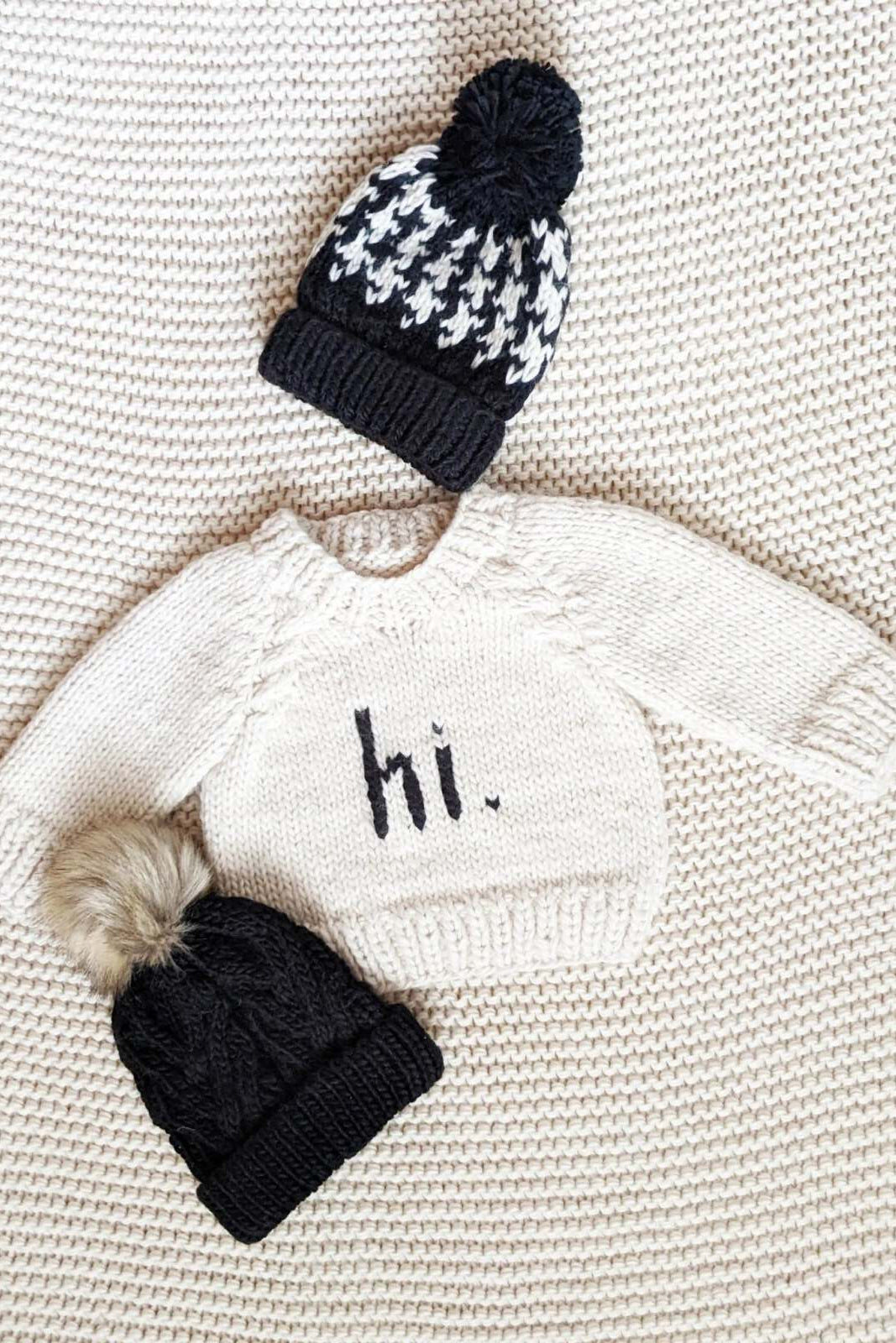 Baby sweater with "hi." text and two knitted hats on a textured background.