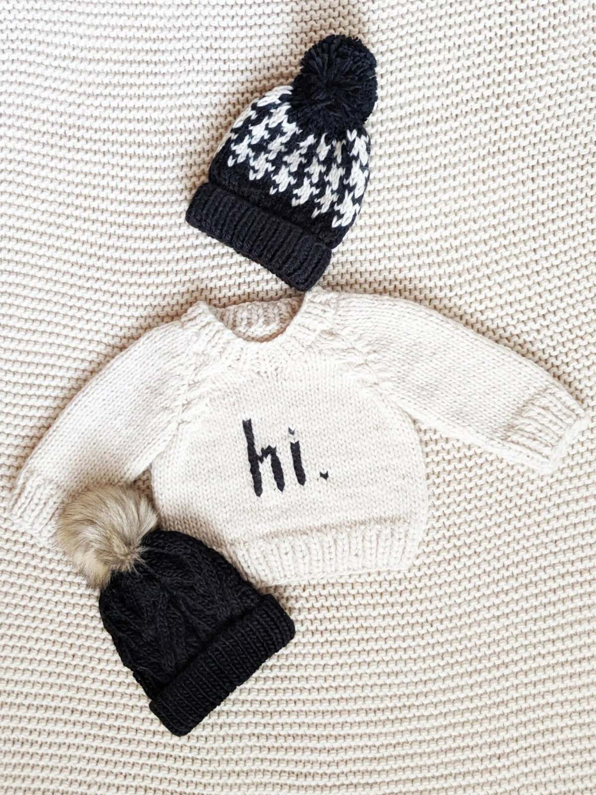 Baby sweater with "hi." text and two knitted hats on a textured background.