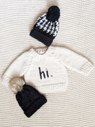 Baby sweater with "hi." text and two knitted hats on a textured background.