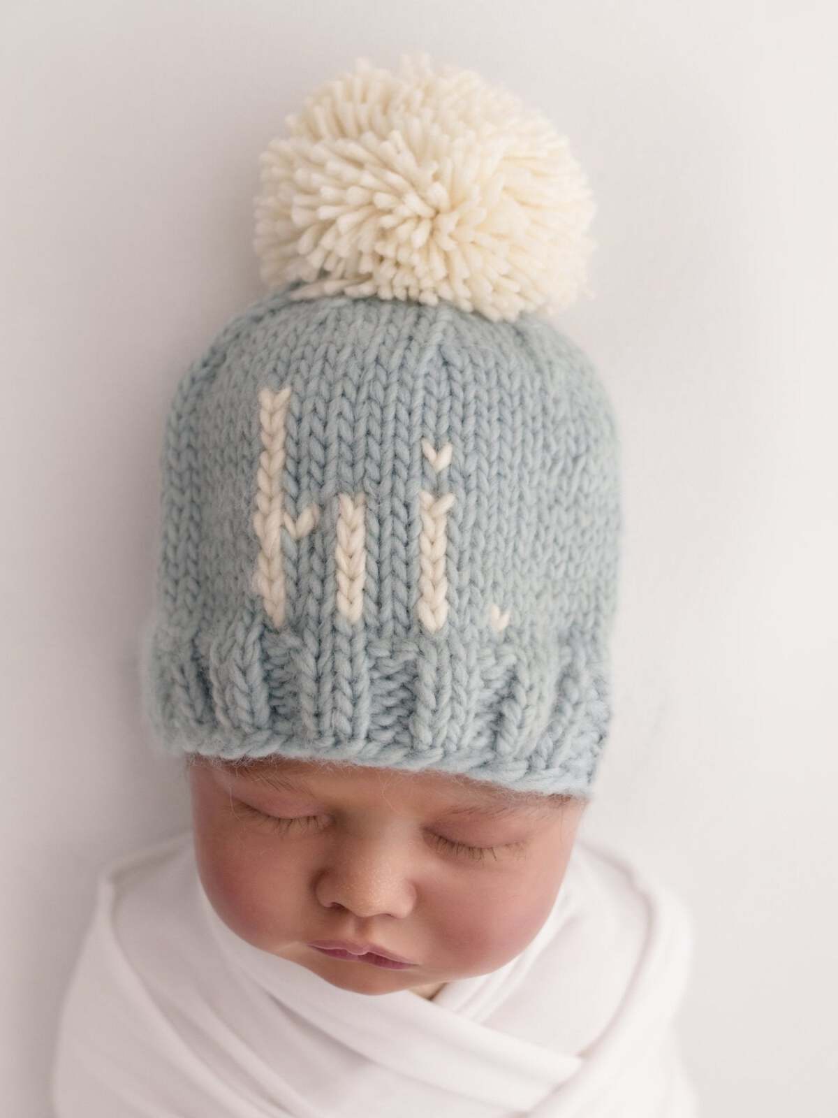 Infant wearing a light blue knit hat with a white pompom and the word "hi" stitched on the front.