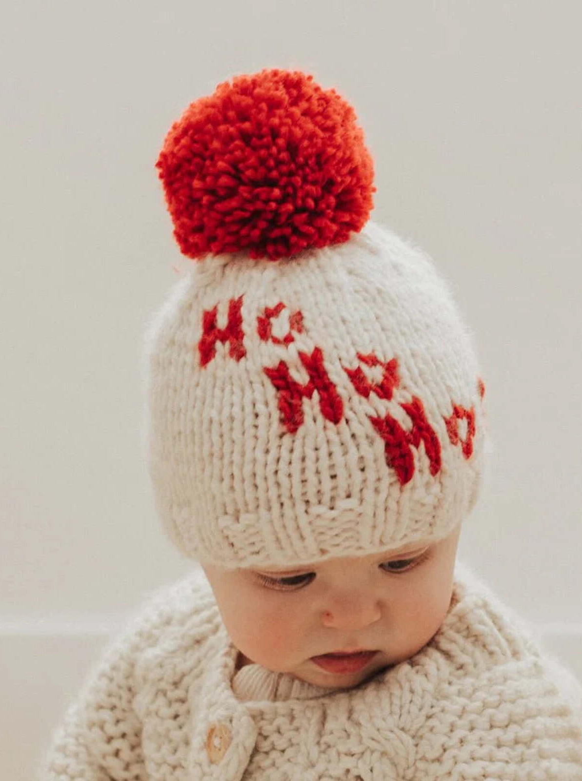 Baby wearing a knitted white hat with a red pom-pom and festive "Ho Ho Ho" design, looking down.