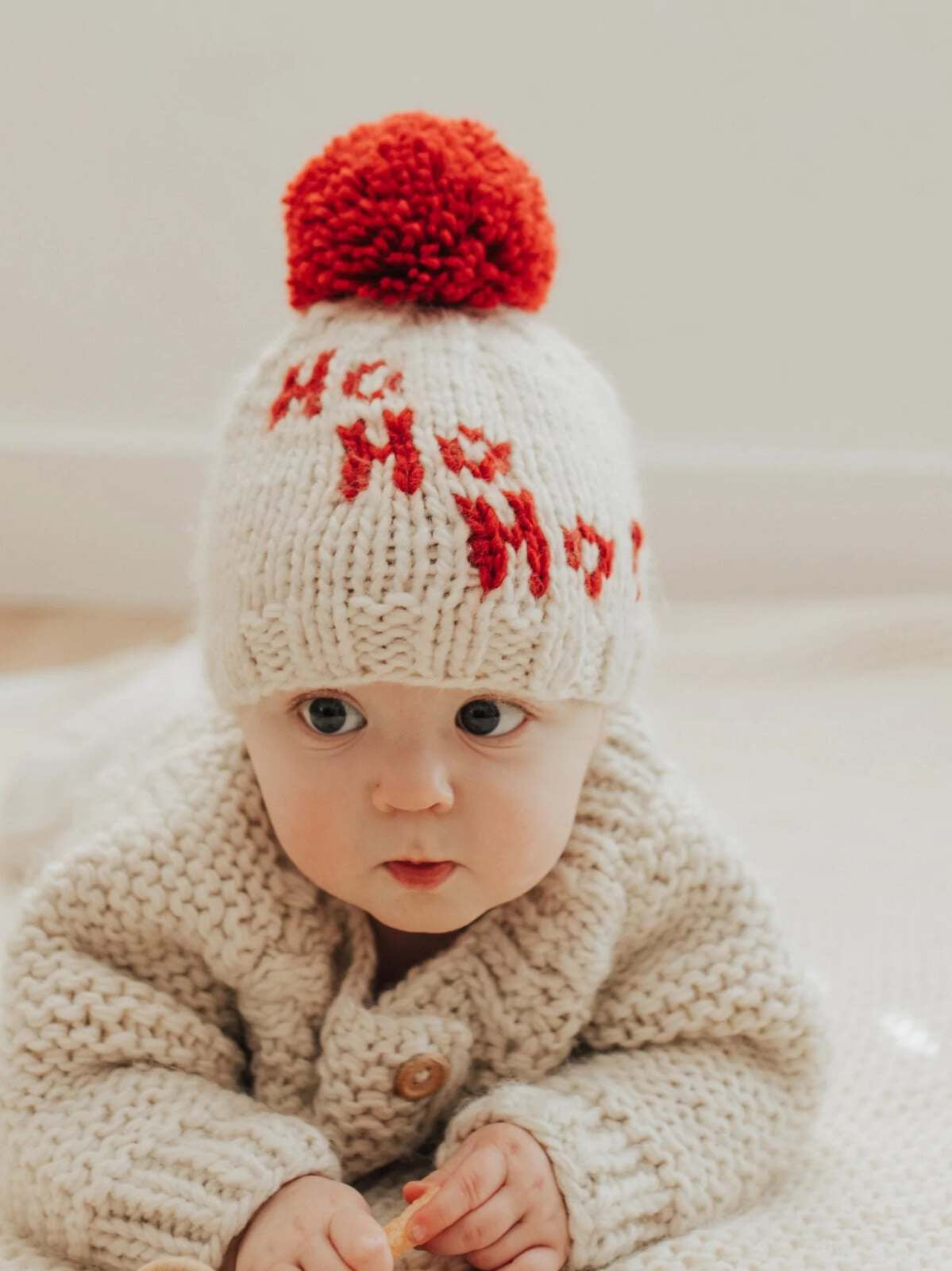 Baby wearing a festive white knit hat with a red pom-pom, sitting on a soft surface, looking curiously at the camera.