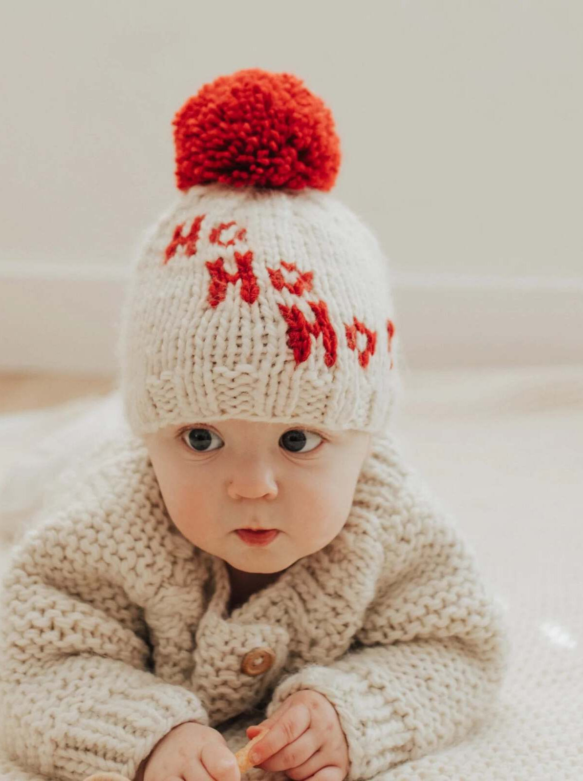 Baby wearing a festive white knit hat with a red pom-pom, sitting on a soft surface, looking curiously at the camera.