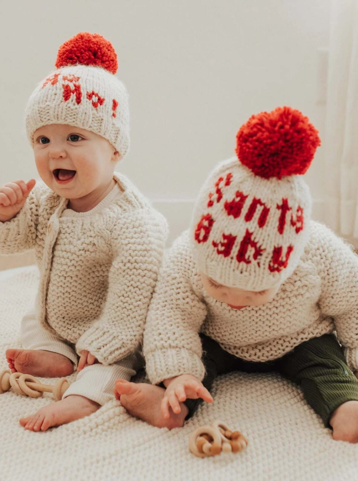 Two smiling babies in knitted sweaters and festive hats, playing on a soft, textured surface.