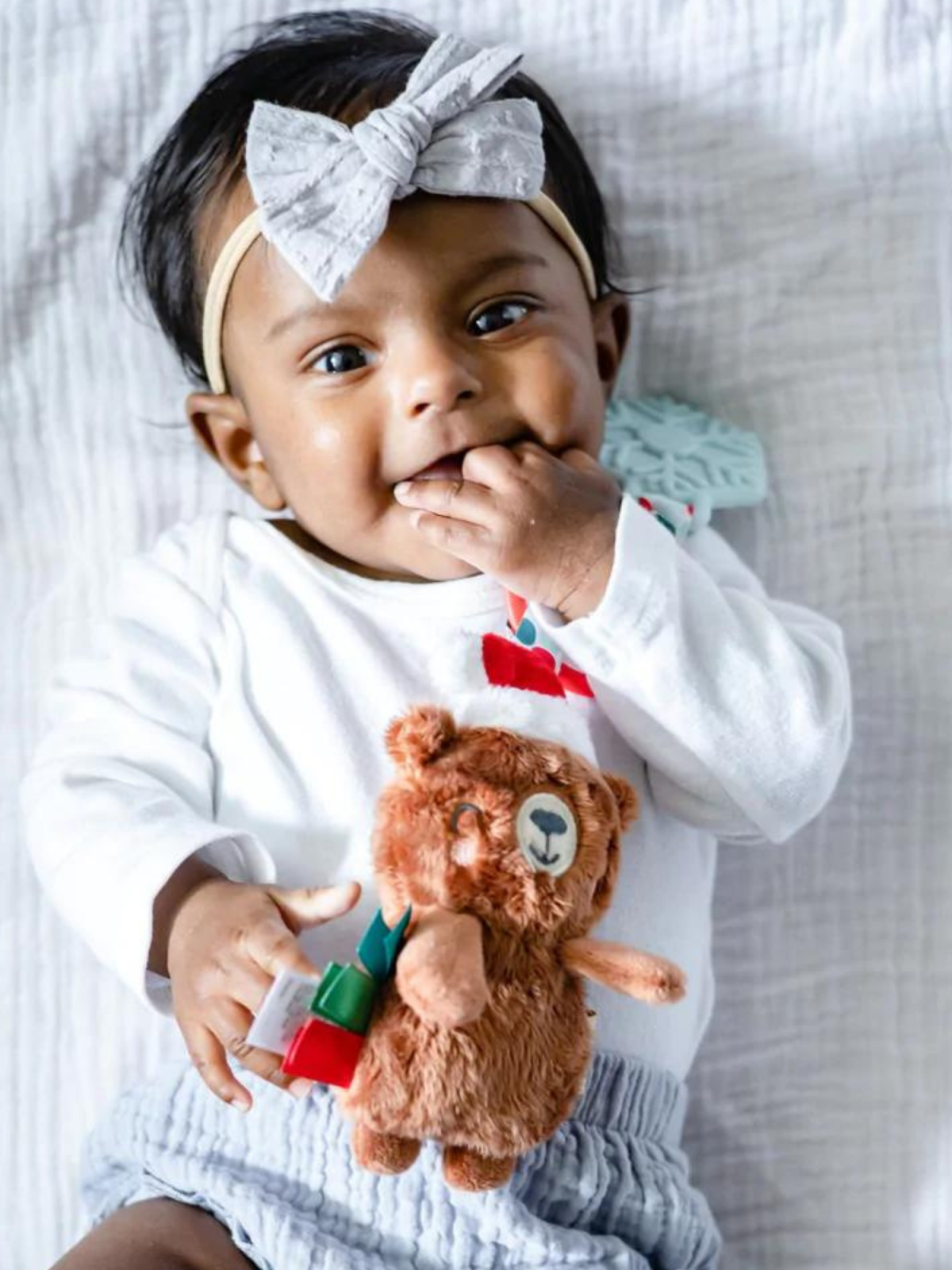 Smiling baby girl with a gray bow, holding a plush bear toy while lying on a blanket.