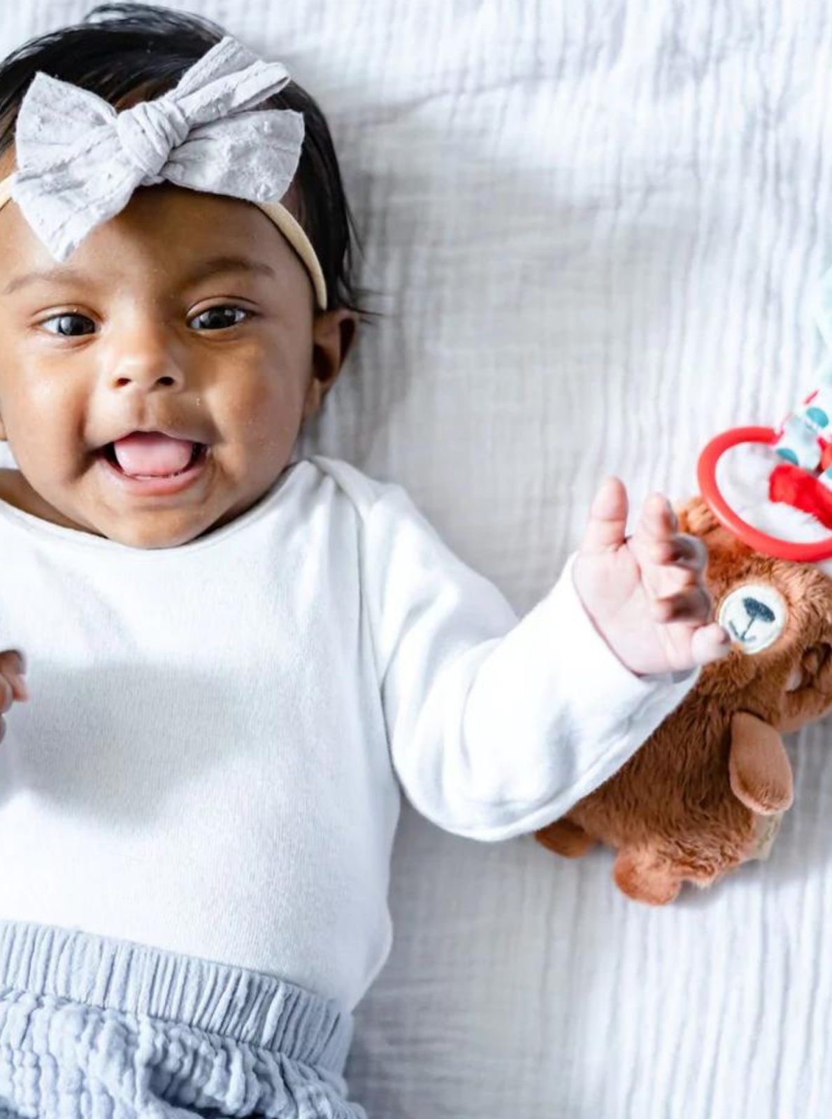 Baby girl with a bow smiles while lying on a blanket, holding a plush toy beside her.