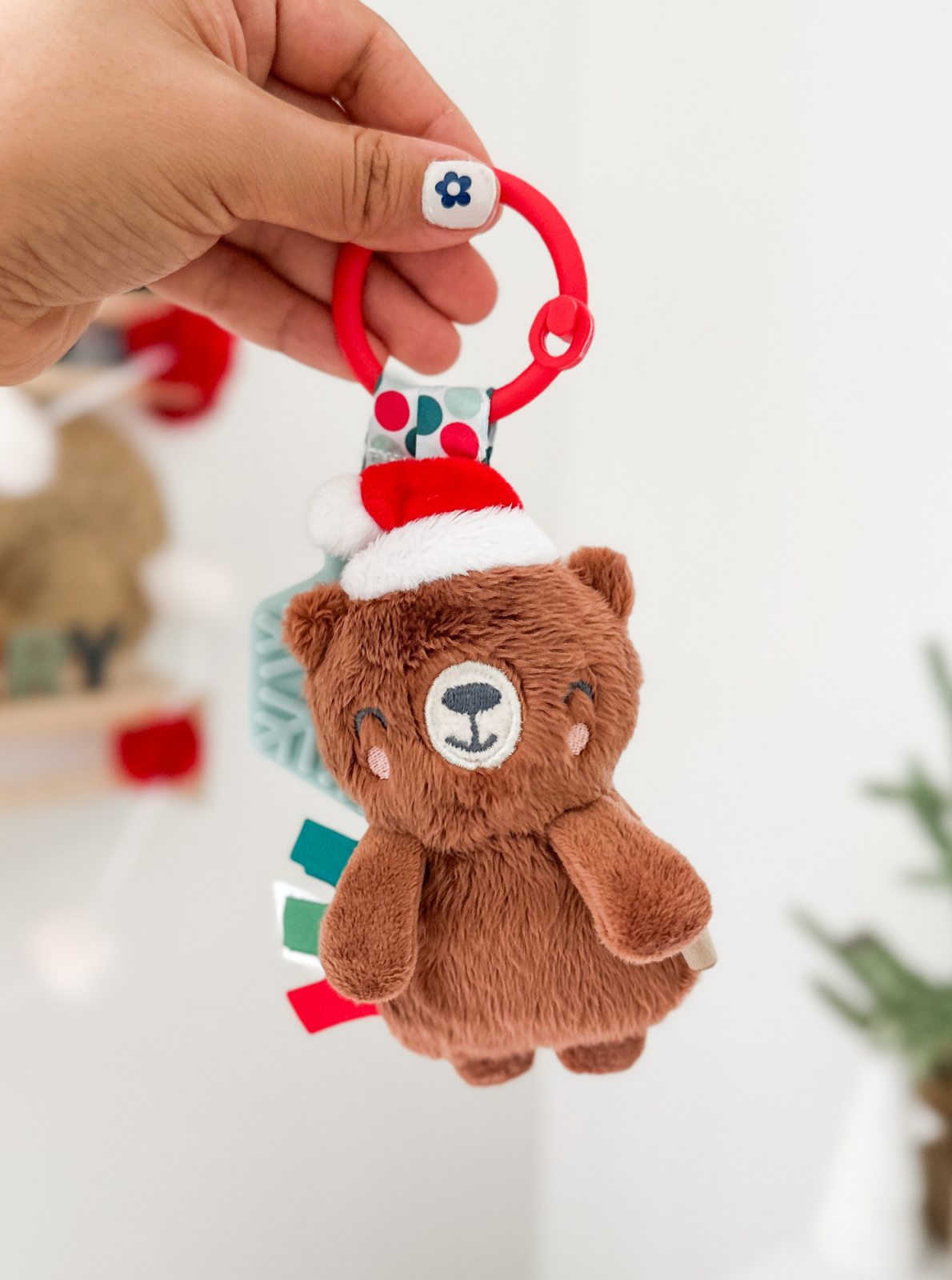 Brown bear plush toy with a festive Santa hat, held by a hand with a flower nail design, against a bright background.