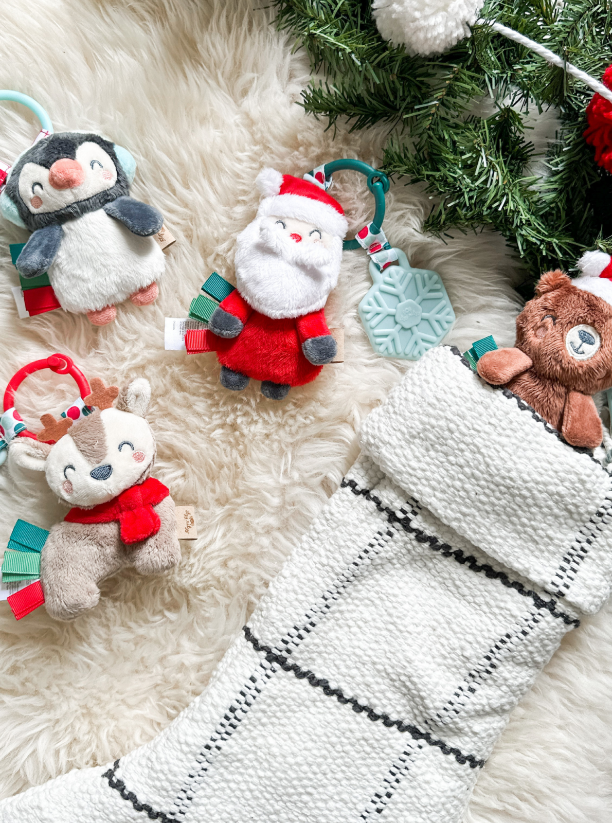 Colorful holiday plush toys near a stocking on a soft, fluffy surface with greenery in the background.