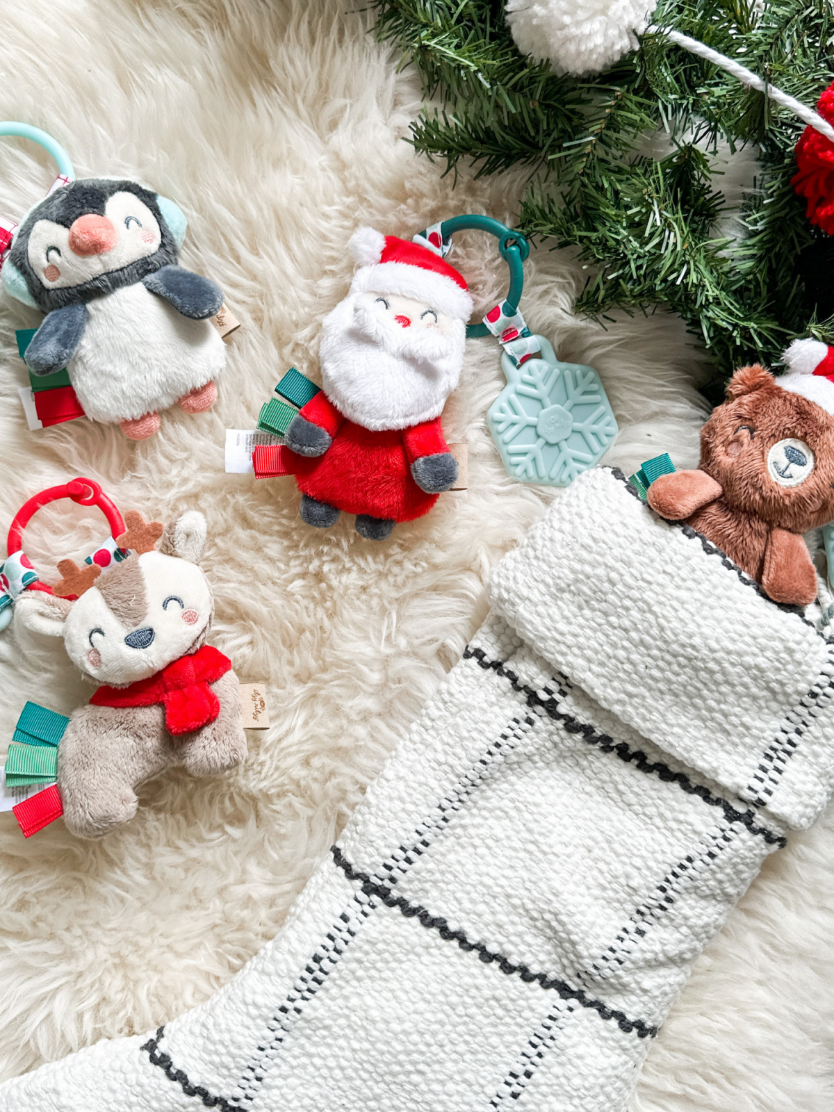 Colorful holiday plush toys near a stocking on a soft, fluffy surface with greenery in the background.