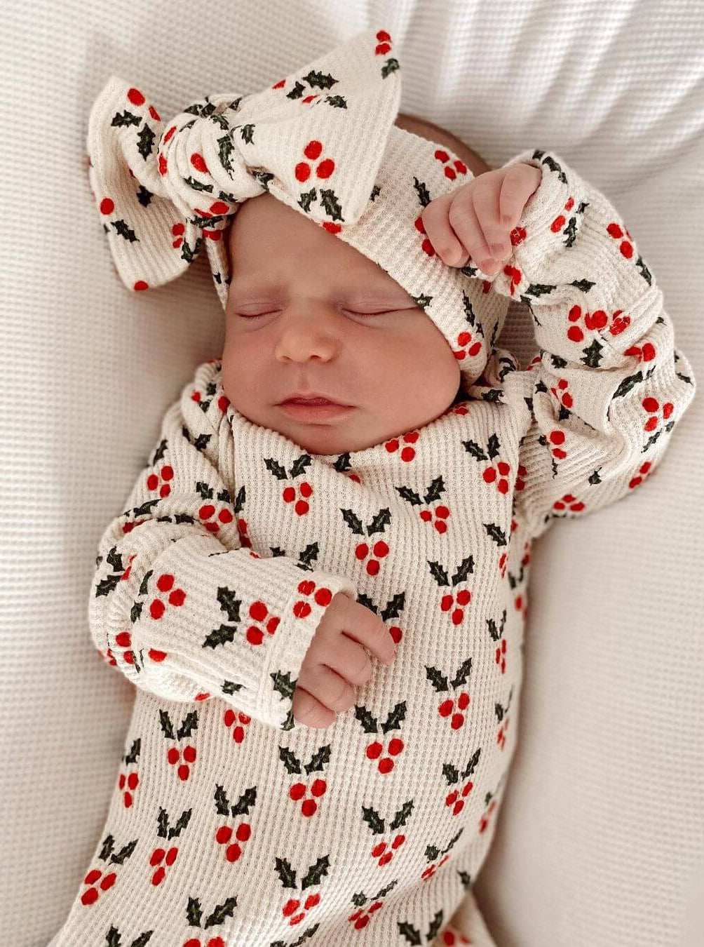 Newborn baby sleeping, wearing a festive outfit with red berries and holly pattern, topped with a bow headband.