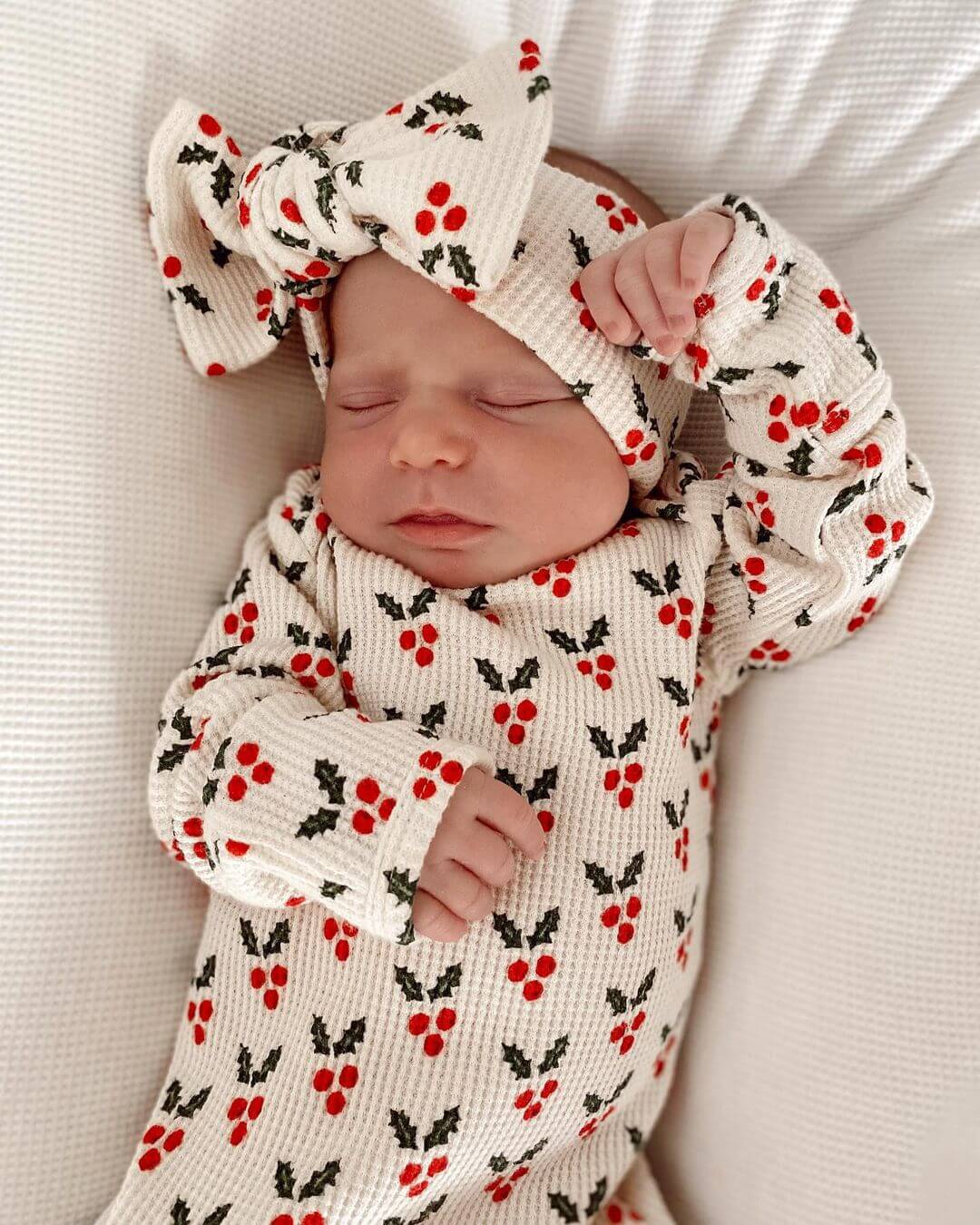 Newborn baby sleeping, wearing a festive outfit with red berries and holly pattern, topped with a bow headband.