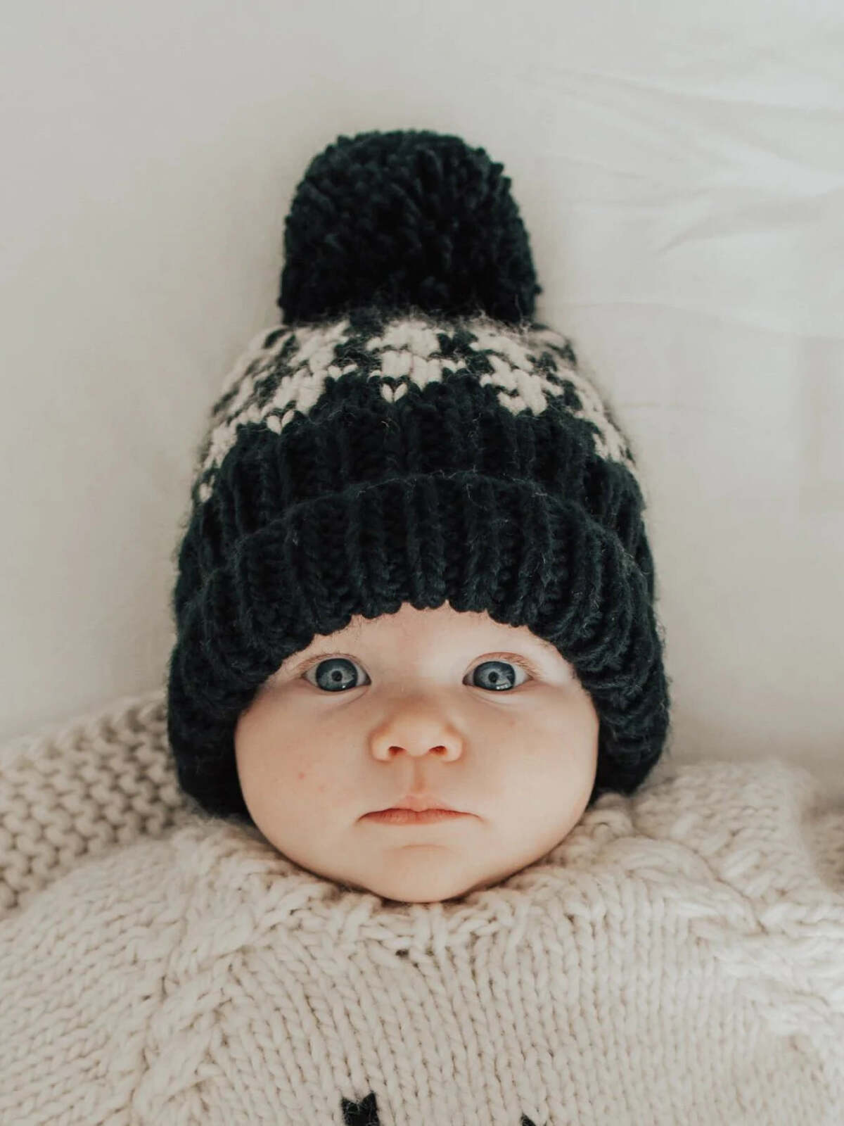 Baby wearing a knitted hat with a pom-pom, wrapped in a cozy blanket, with big blue eyes looking up.
