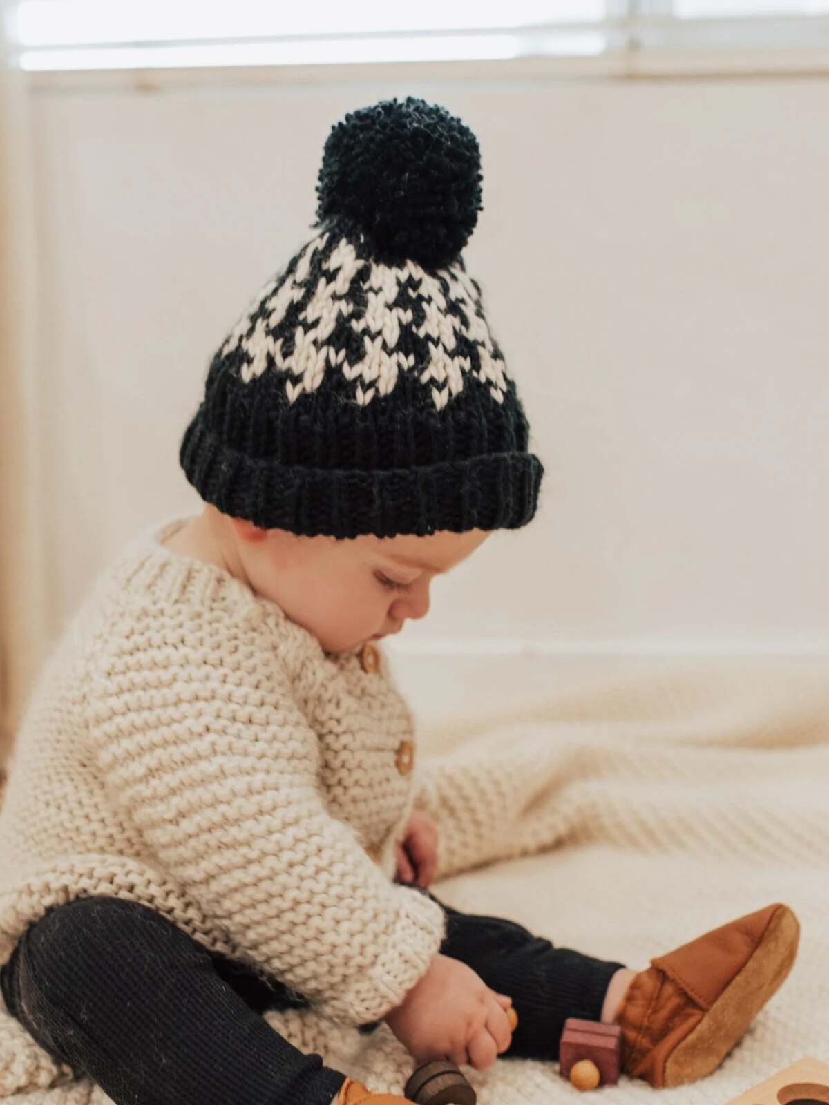 Baby in a cozy knitted sweater and a black-and-white pom-pom beanie, playing on a soft blanket.