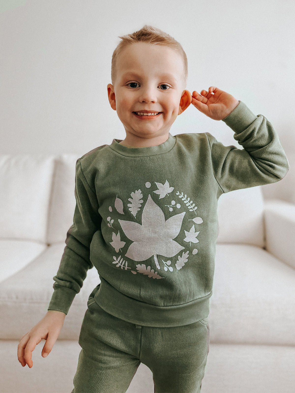 Smiling child in green outfit with leaf design, standing on a sofa, waving hand cheerfully.