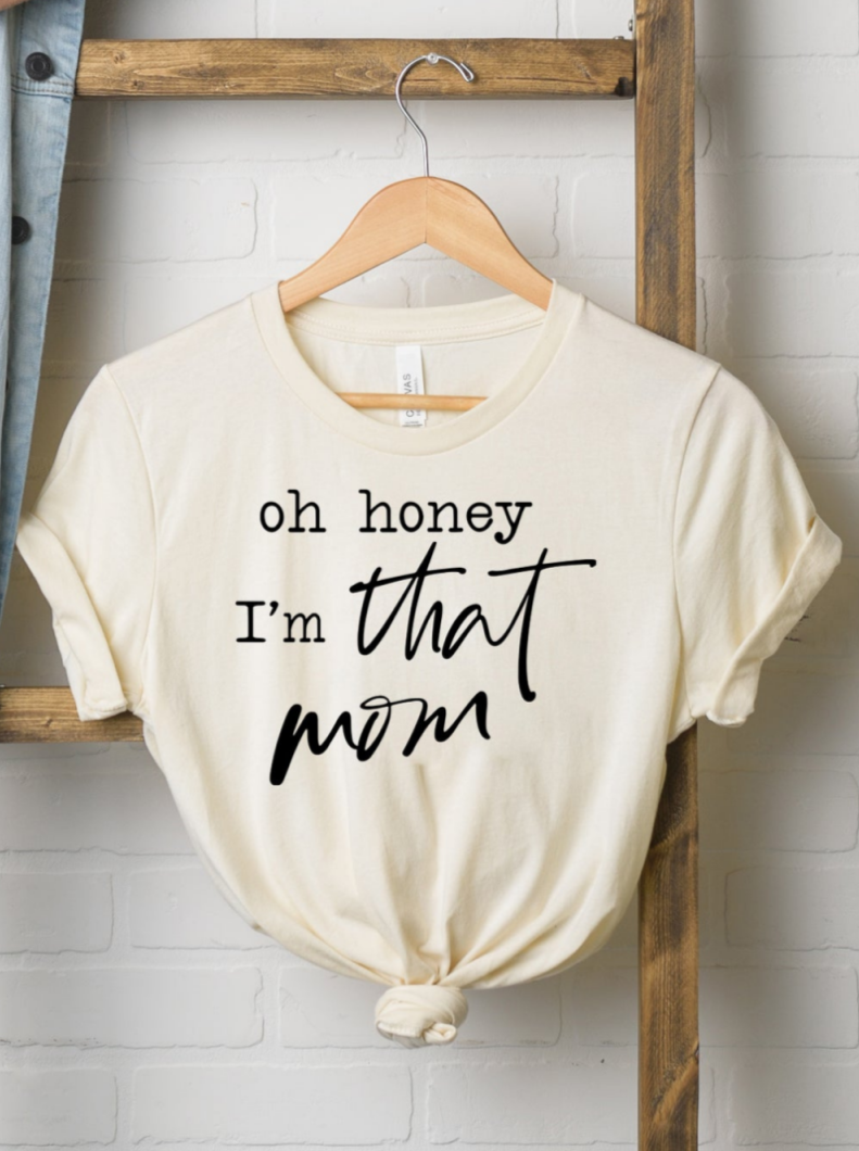 Cream t-shirt hanging on a wooden hanger with the text "oh honey I'm that mom" printed in bold black letters.