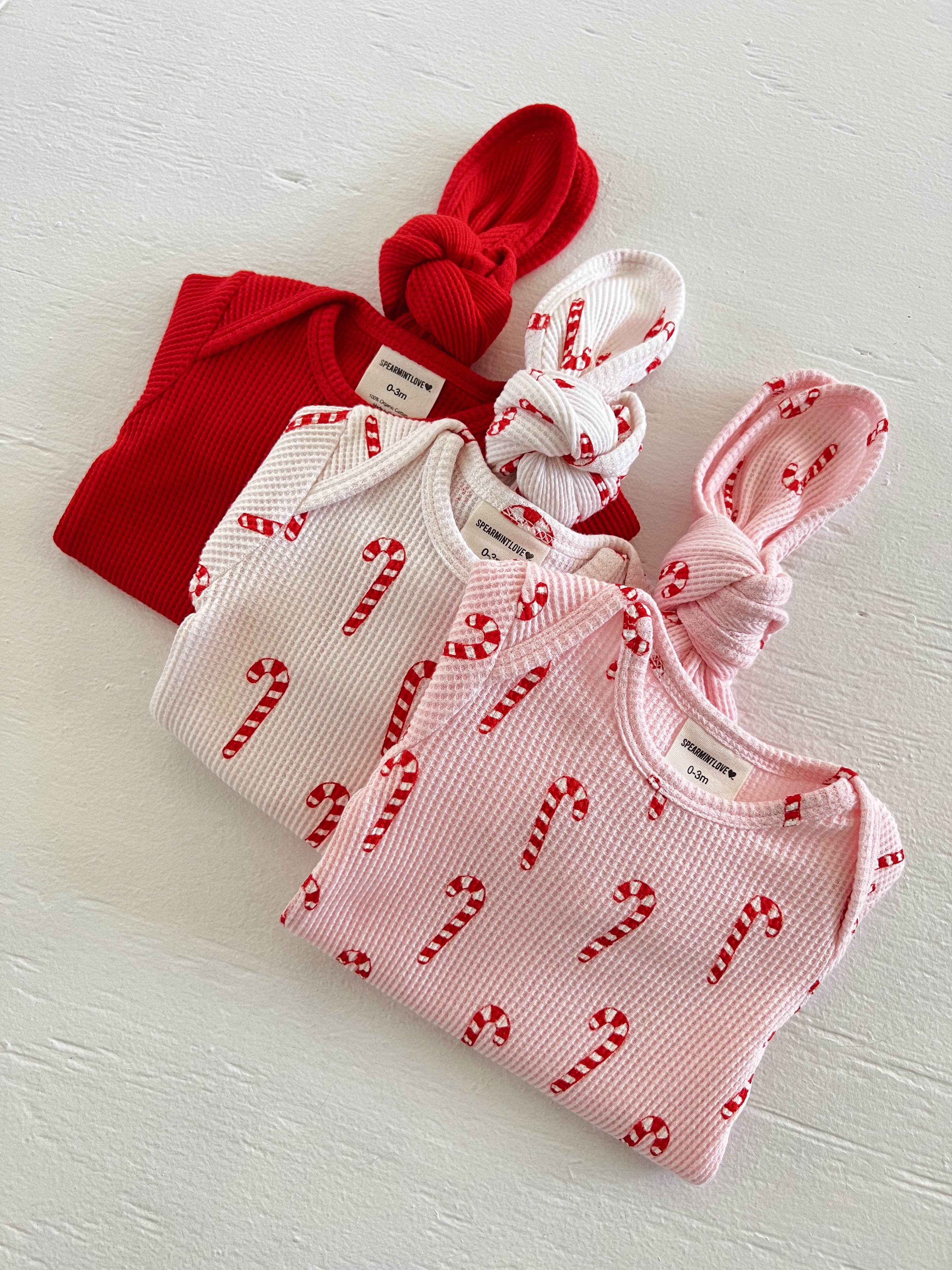 Three baby outfits in red, white, and pink with candy cane patterns, featuring knotted tops and soft fabric.