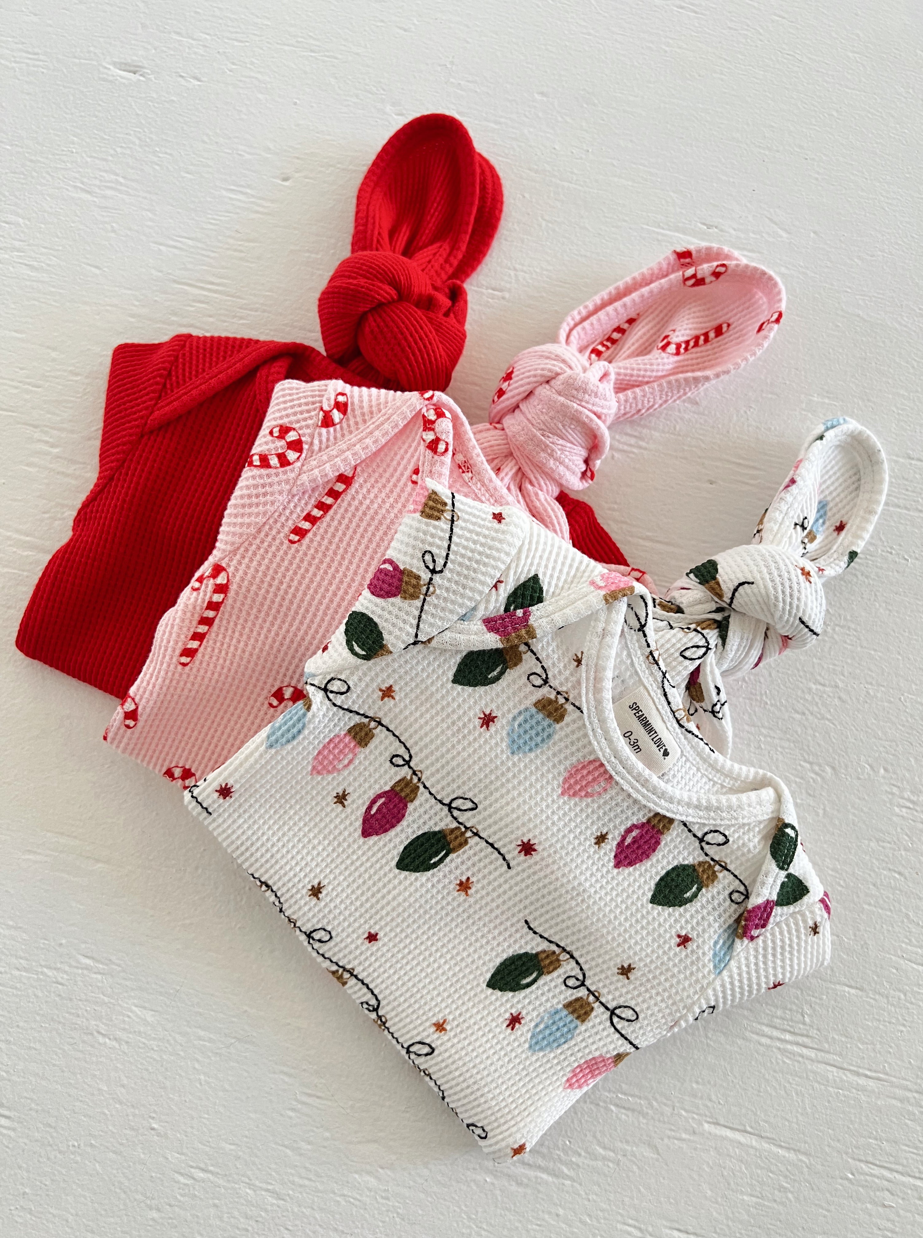 Three toddler hats in red, pink candy cane pattern, and white with holiday lights on a textured surface.