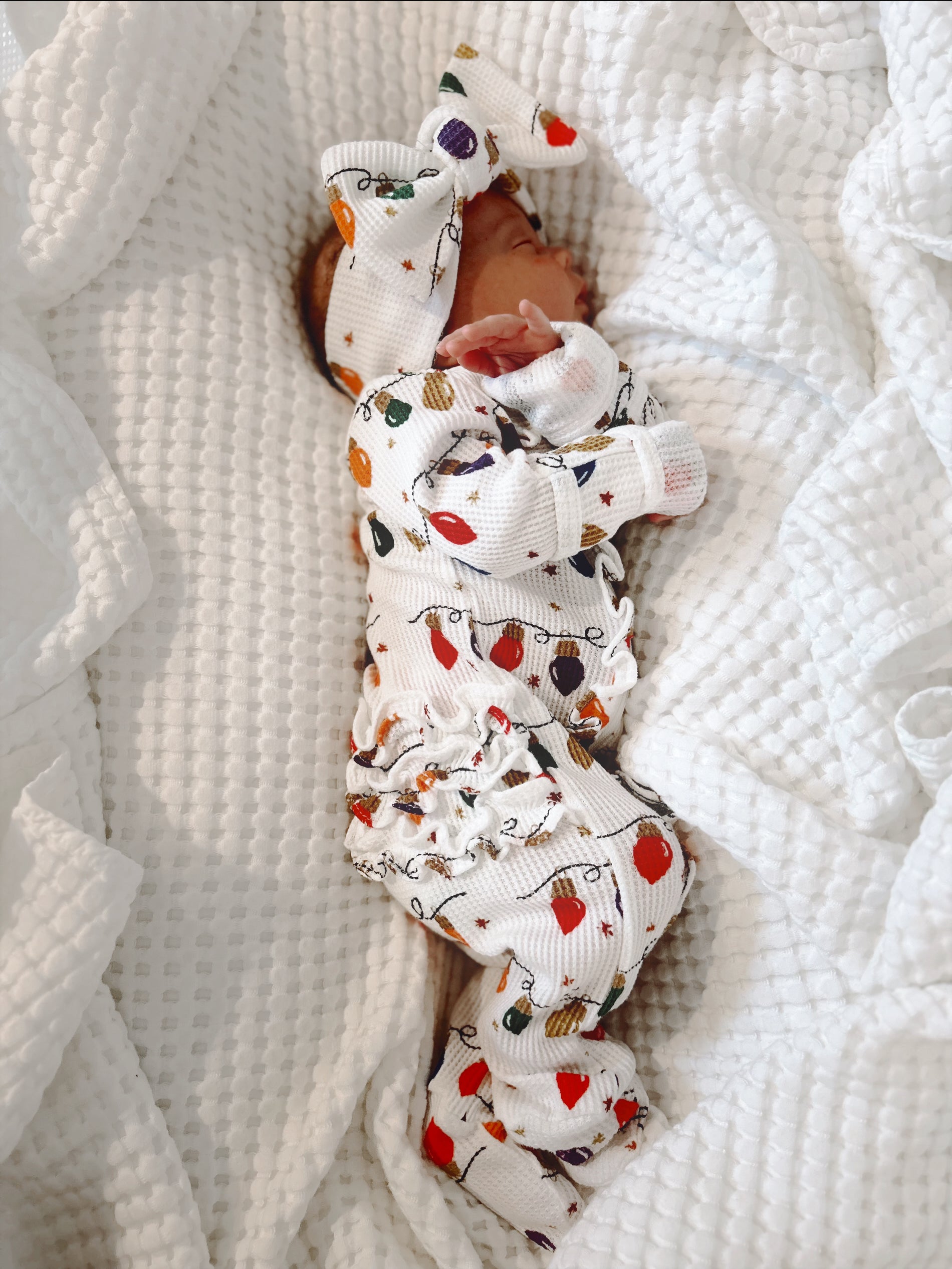 Newborn wearing festive patterned outfit and large bow, peacefully sleeping on white blanket.