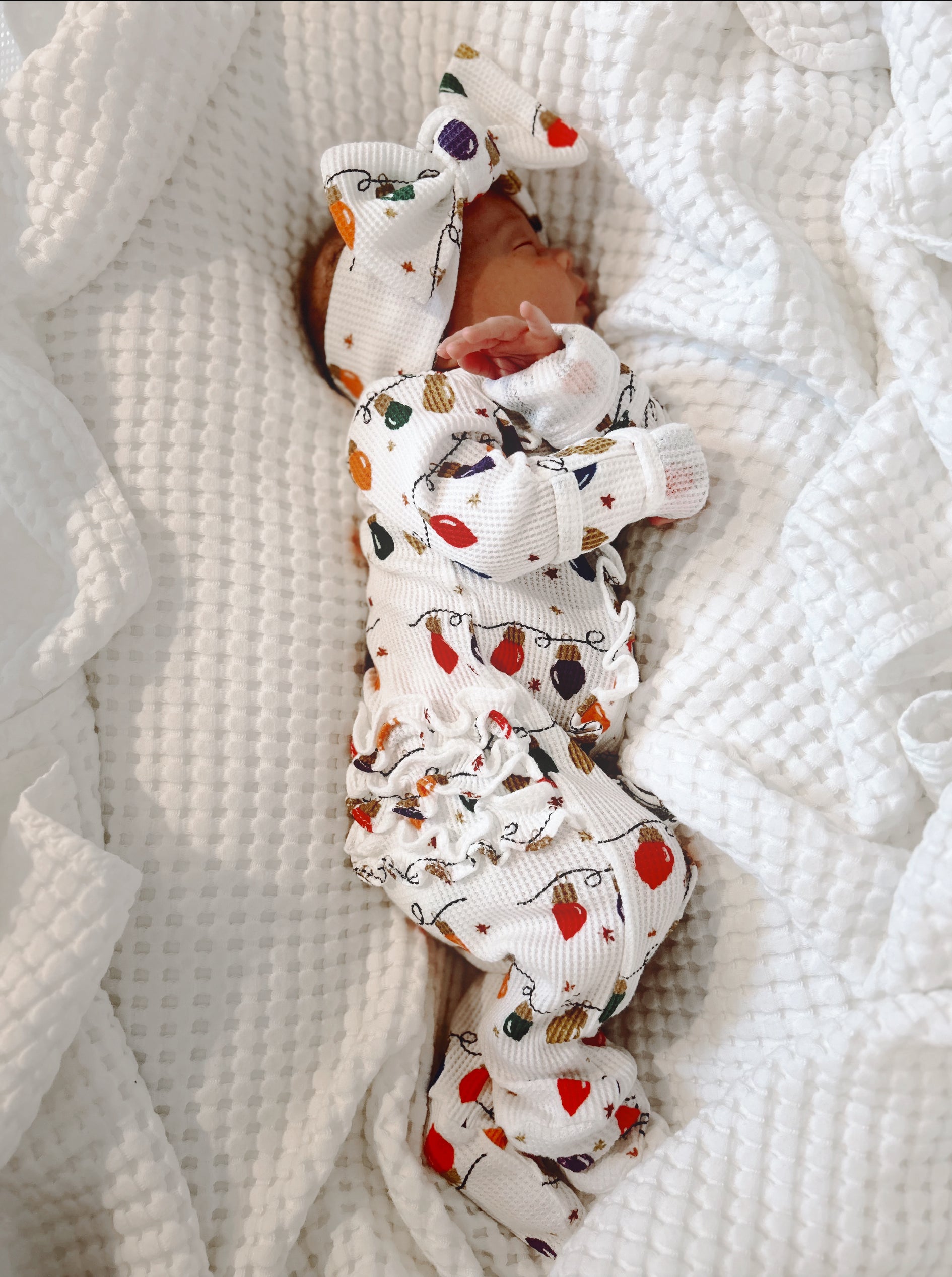 Newborn wearing festive patterned outfit and large bow, peacefully sleeping on white blanket.