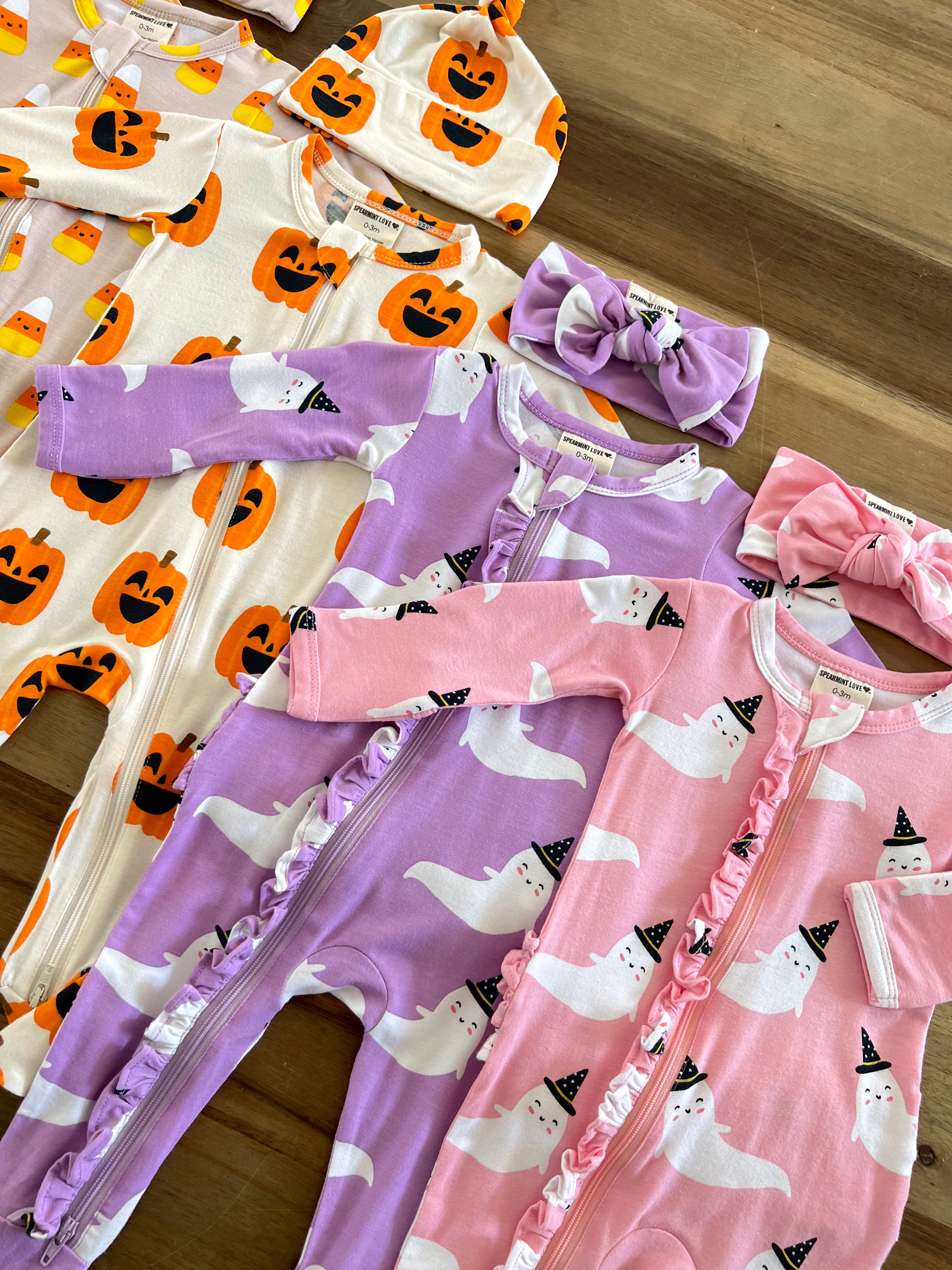 Colorful baby onesies with Halloween themes: pumpkins, ghosts, and matching accessories on a wooden surface.