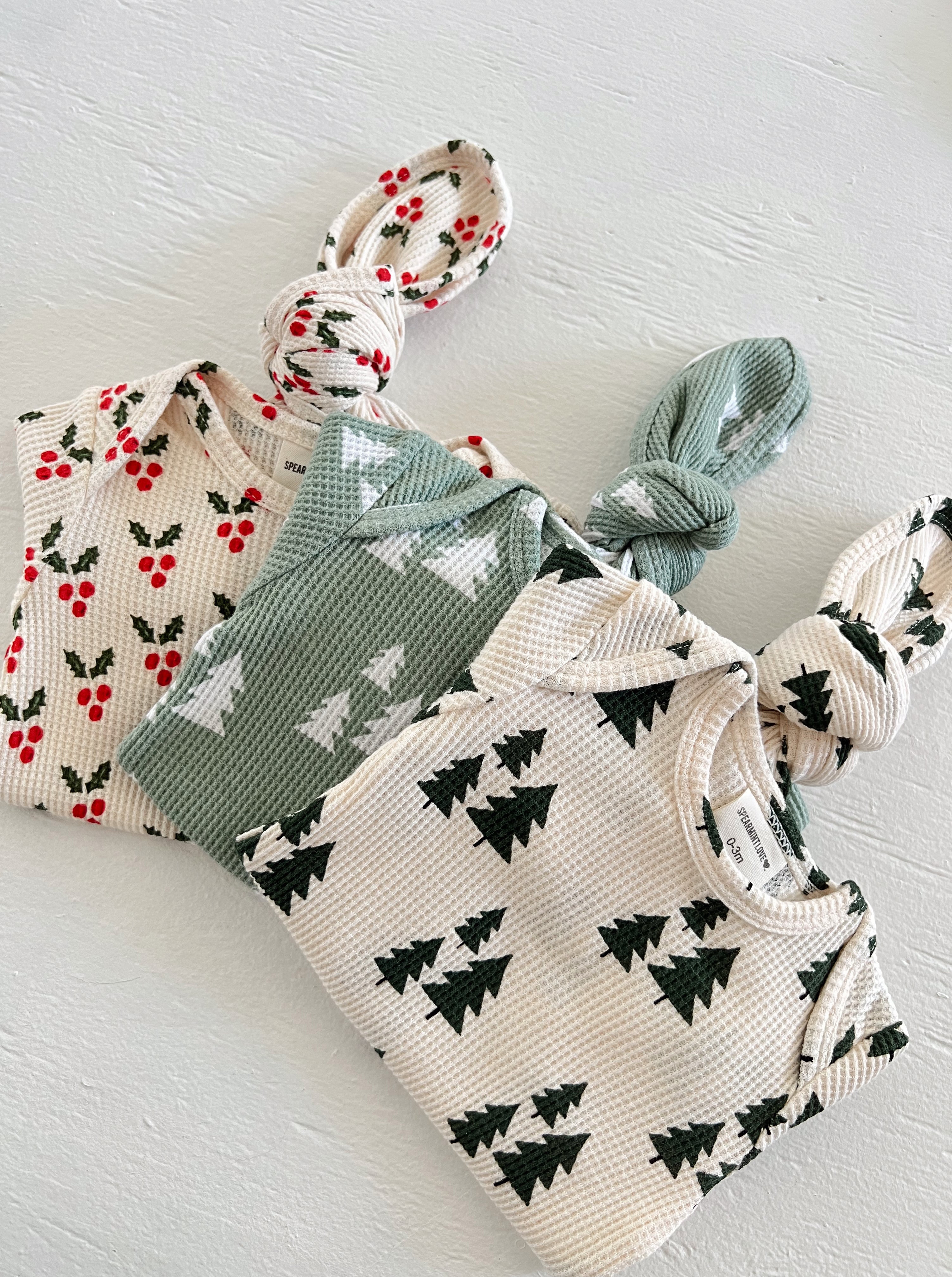 Three patterned baby outfits: one with red berries, one with green trees, and one with a knot at the top.