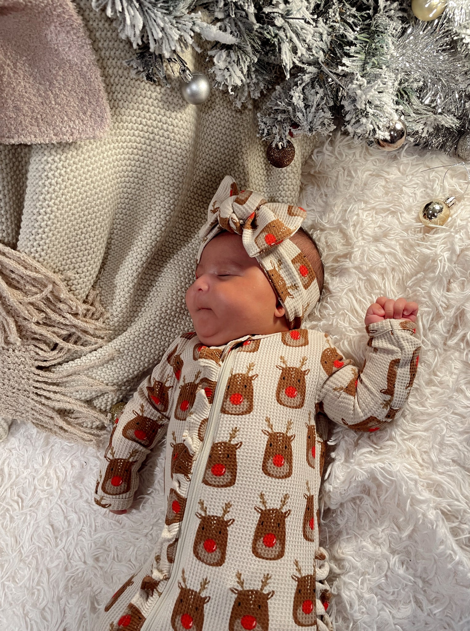 Baby in a festive reindeer outfit resting on a cozy blanket near a decorated tree.