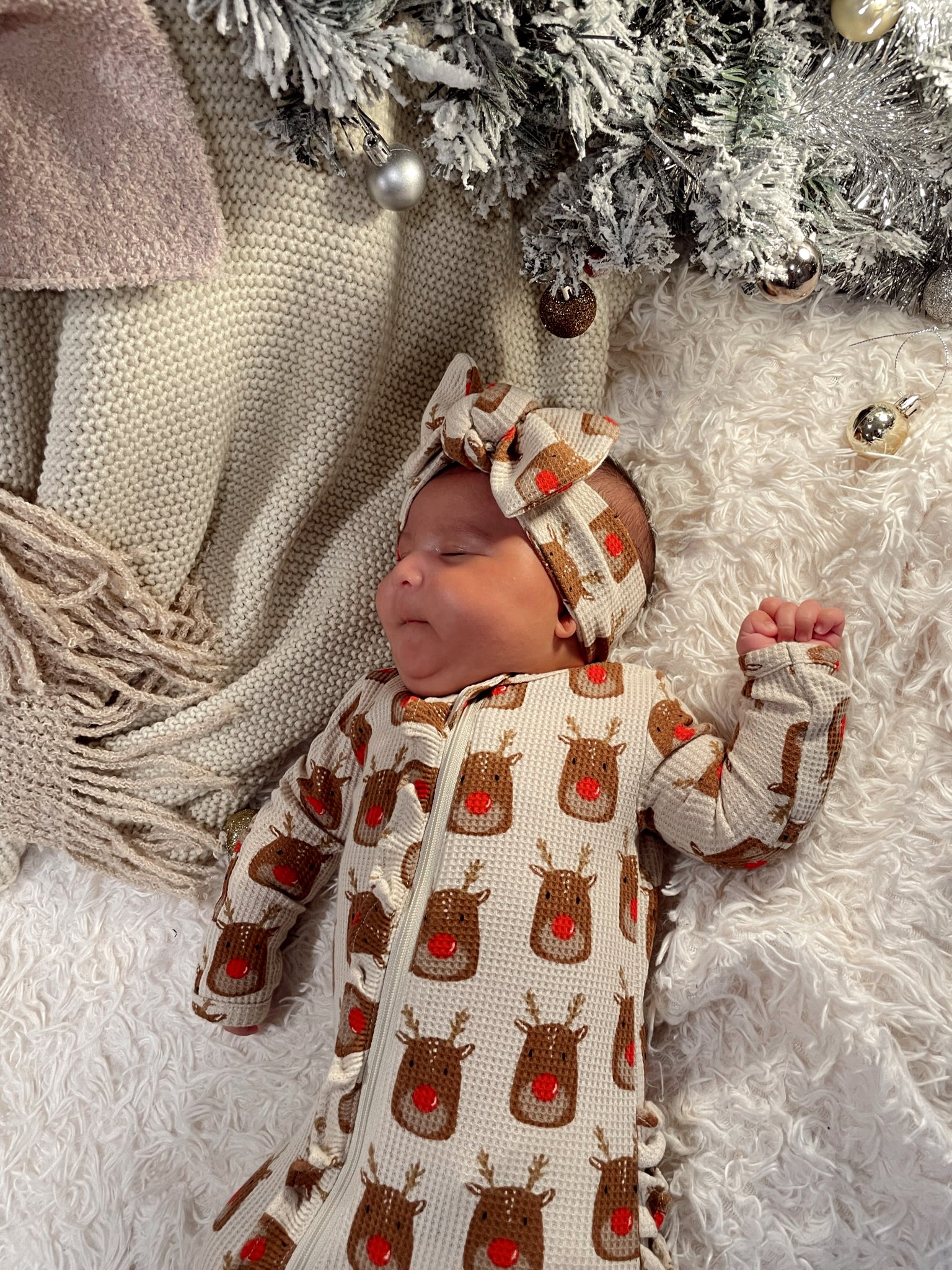 Baby in a festive reindeer outfit resting on a cozy blanket near a decorated tree.