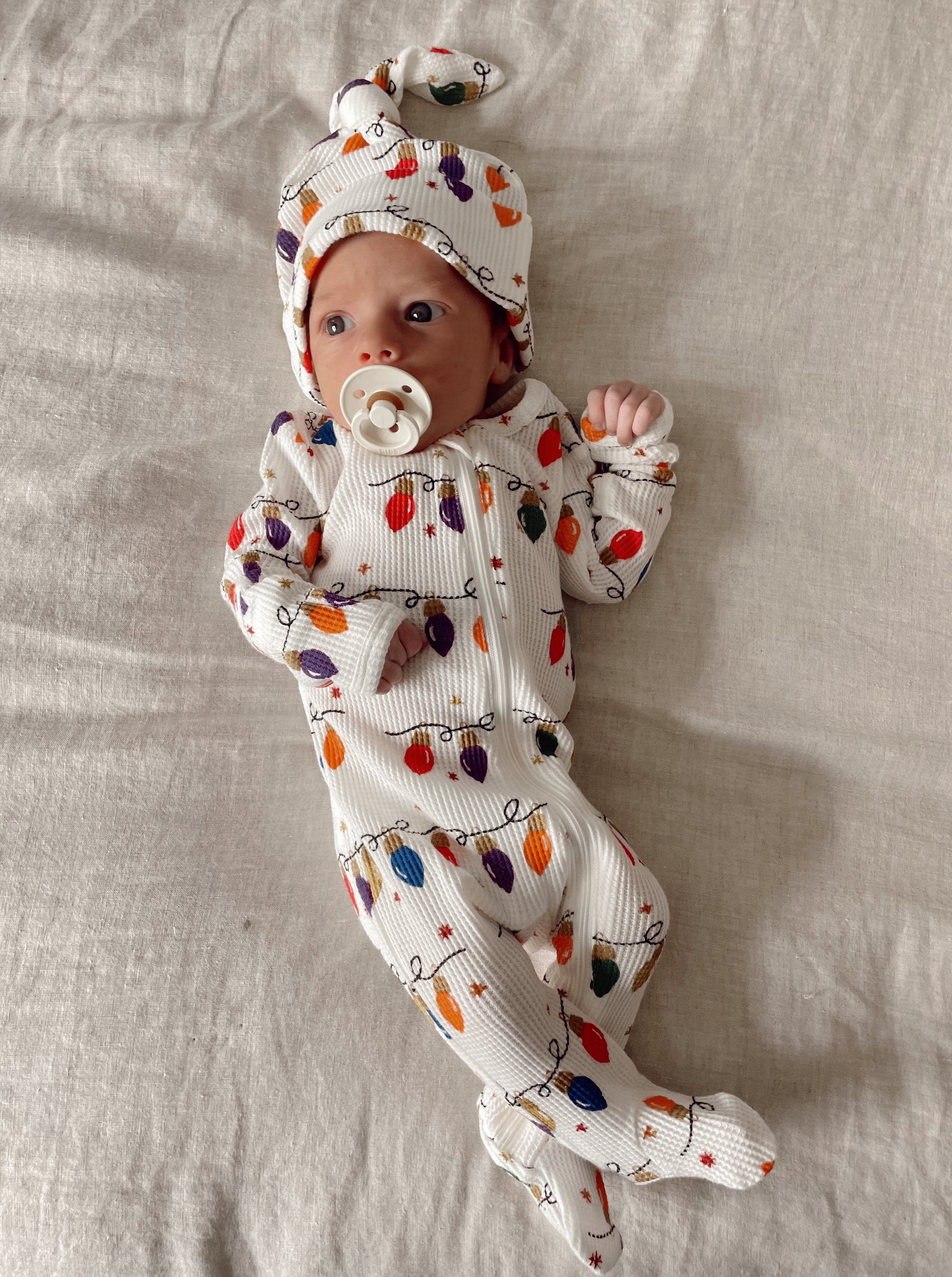 Newborn baby in festive outfit with colorful lights, lying on a light-colored blanket, sucking a pacifier.