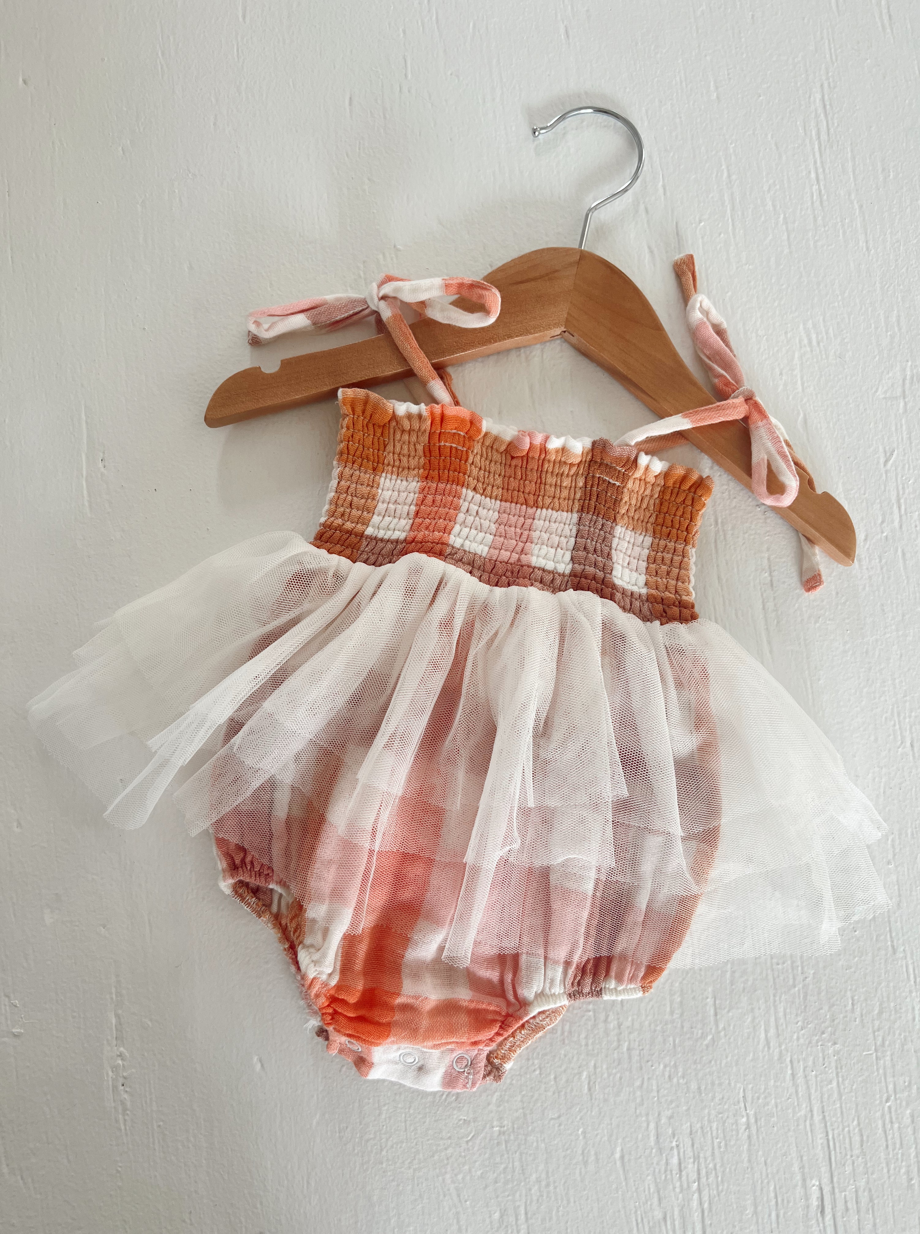 Pink and orange checkered baby romper with tulle skirt, hanging on a wooden hanger against a white wall.