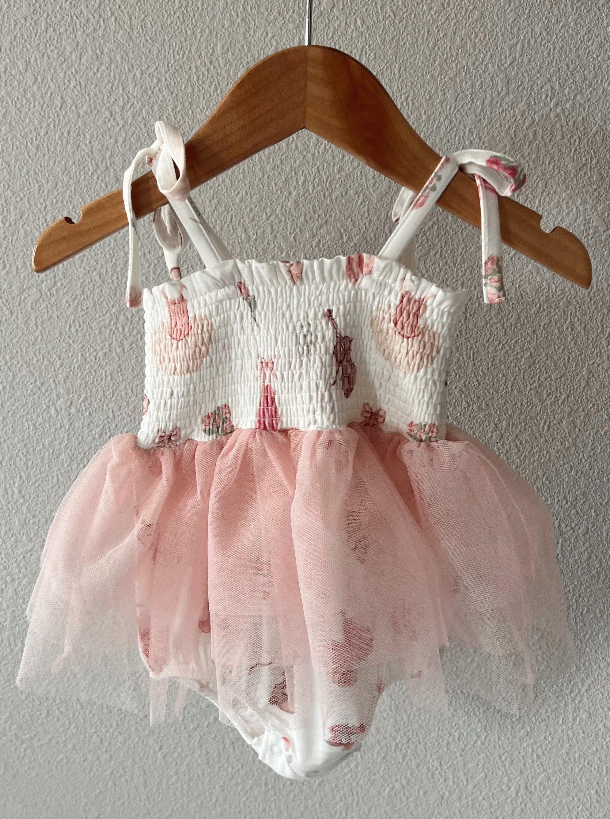 Baby dress in white with floral print and pink tulle skirt, hanging on a wooden hanger against a textured wall.