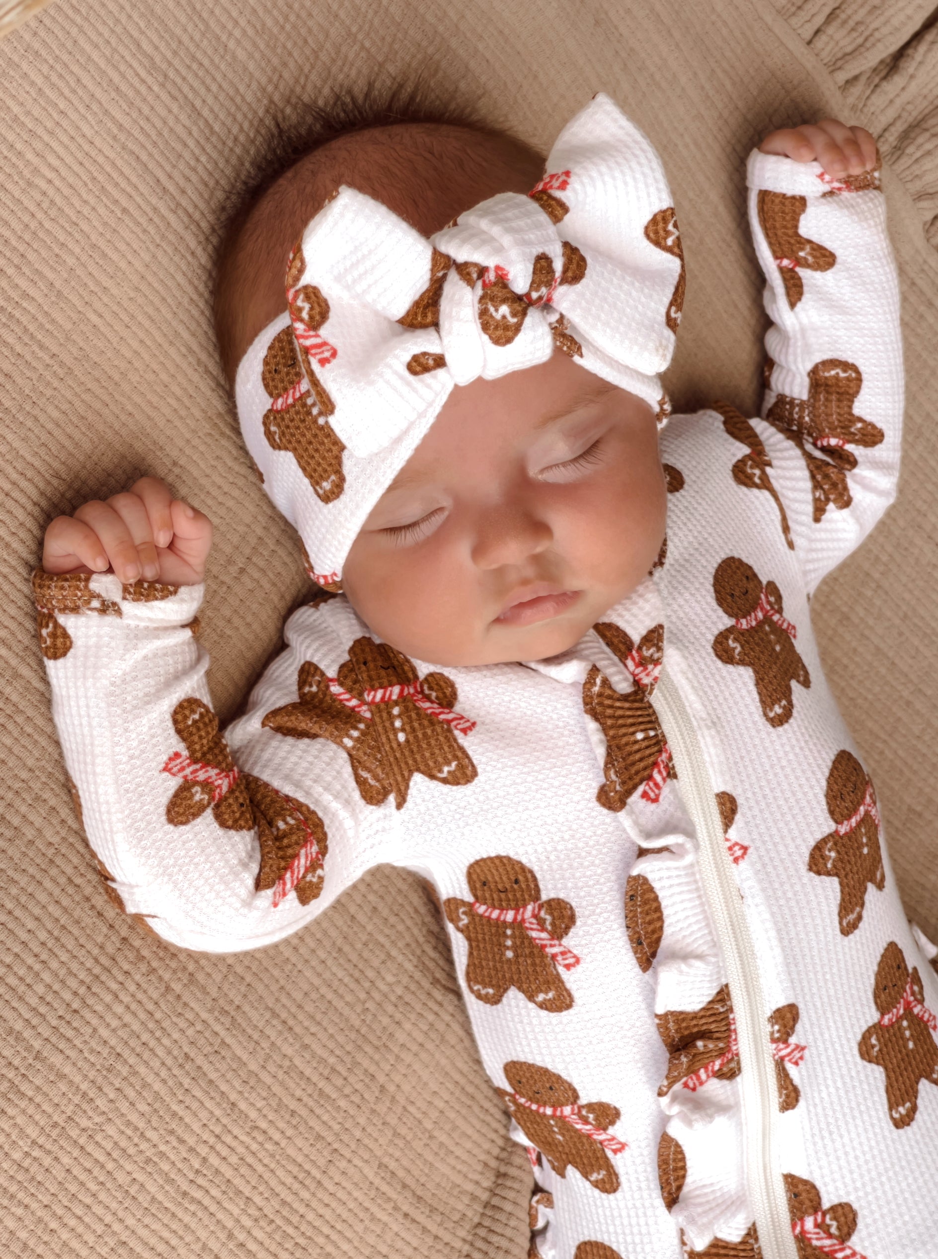 Baby sleeping peacefully in a gingerbread-patterned onesie and headband, resting on a soft surface.